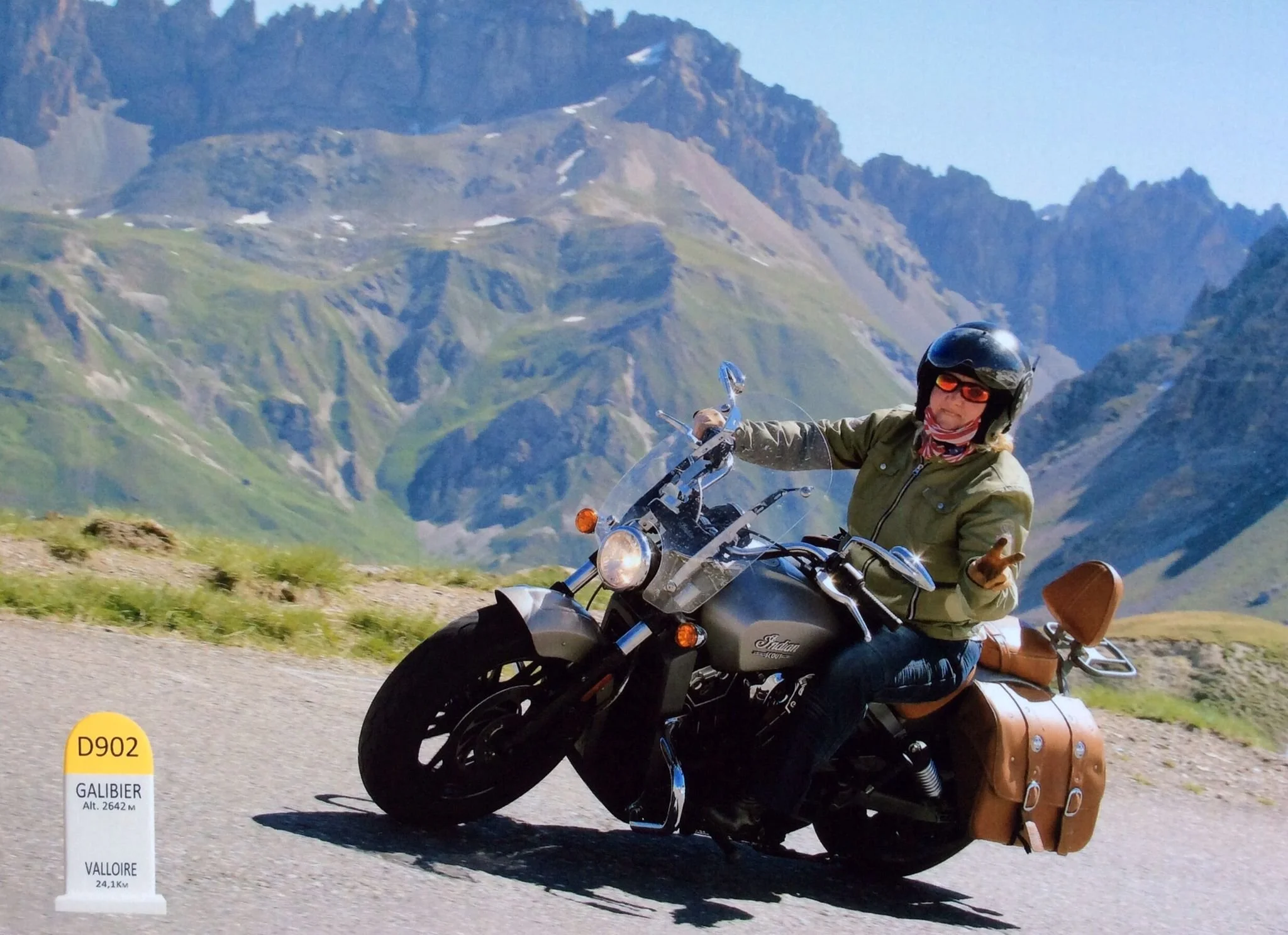 A person riding a black motorcycle on a mountain road with a scenic mountainous background, wearing a helmet and jacket, with a brown leather saddlebag attached to the motorcycle.