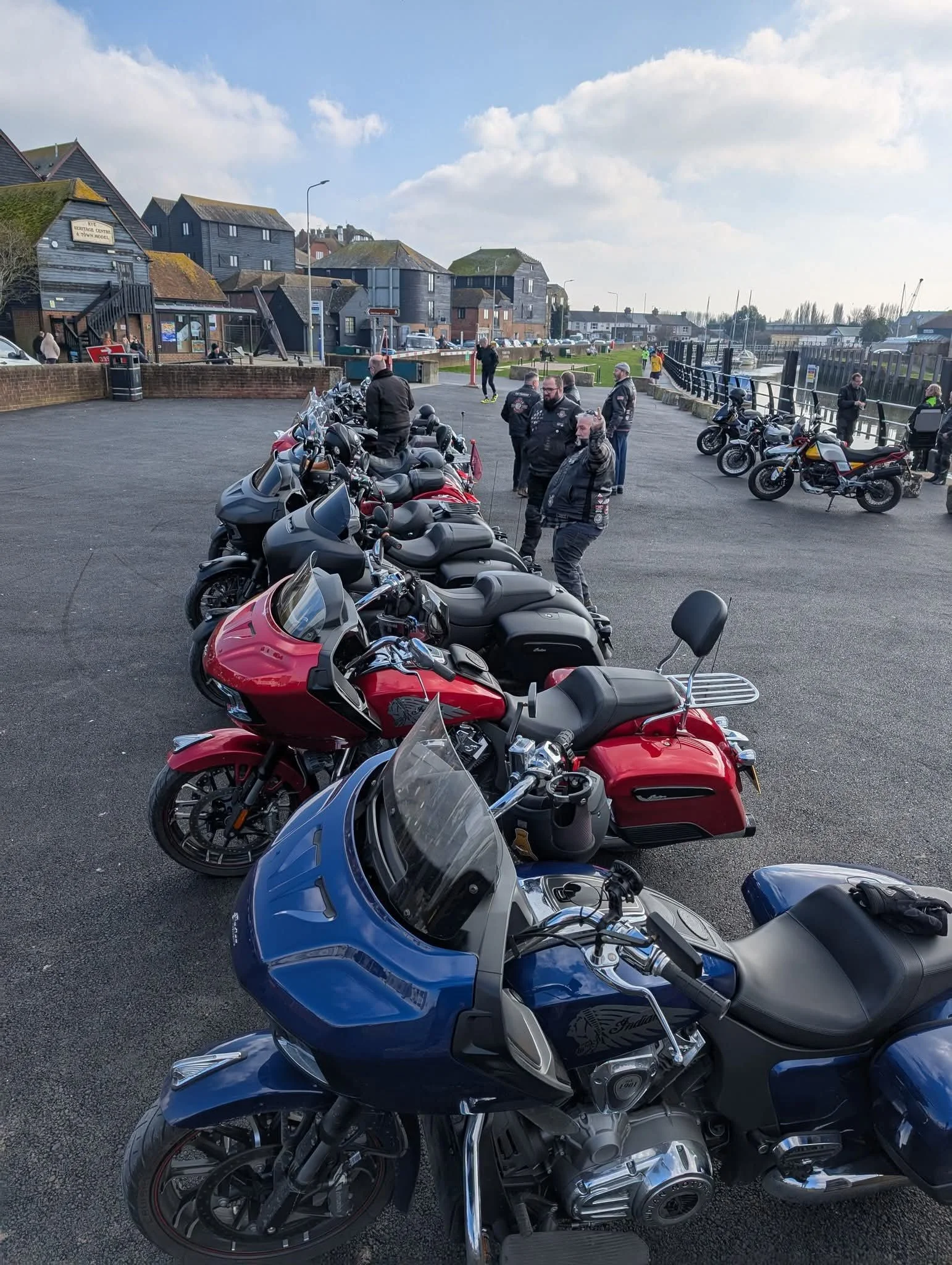 Row of motorcycles parked along a waterfront promenade with people standing and walking nearby, buildings and boats visible in the background under partly cloudy skies.