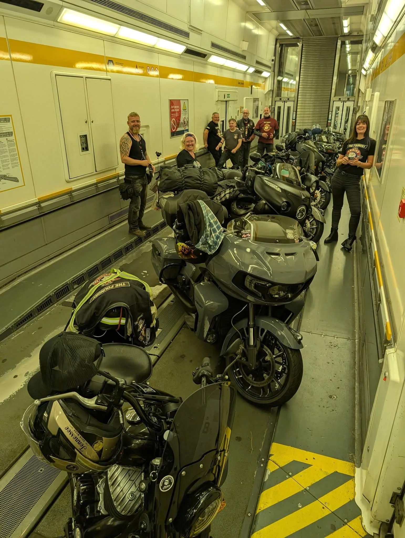 A group of seven people, including four men and two women, are standing inside a vehicle on a ferry with motorcycles parked in front of them. The setting appears to be inside a vehicle ferry.