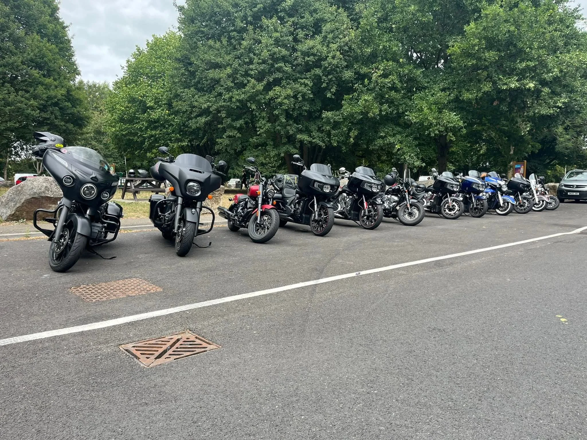 Line of parked motorcycles on a paved parking lot with large green trees in the background.