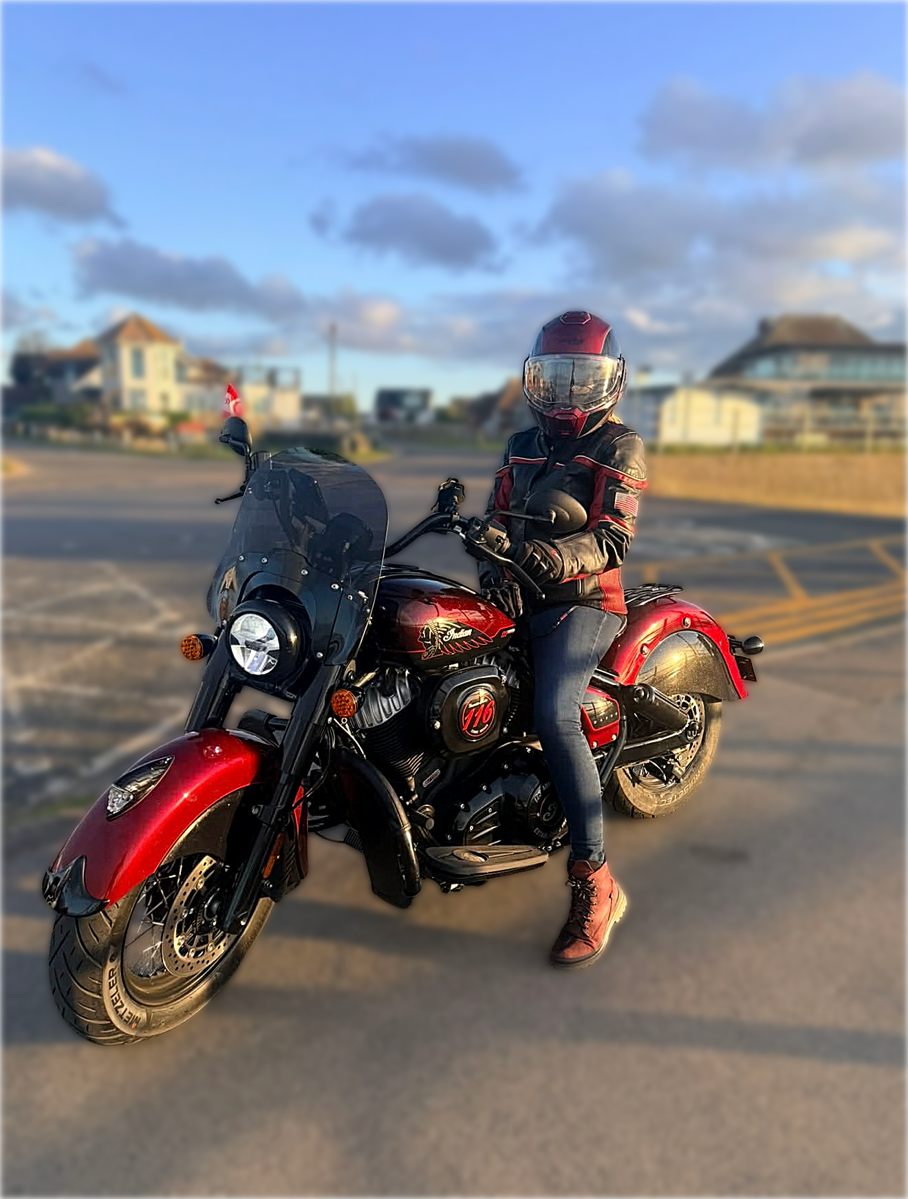 Person wearing a full motorcycle helmet, black and red riding jacket, and jeans, sitting on a red Indian motorcycle in an outdoor parking lot during sunset, with houses and clouds in the background.