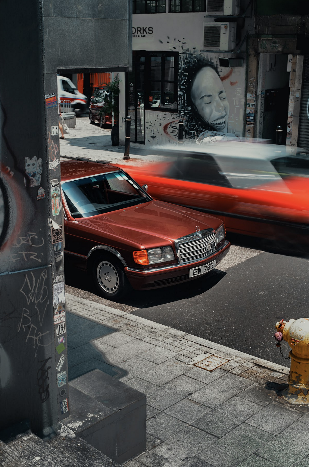 A red vintage Mercedes-Benz parked on city street with a blurred red car passing by at high speed. In the background, there is a building with street art of a smiling woman's face on the wall and graffiti on other walls. The scene includes sidewalk, fire hydrant, and other urban elements.