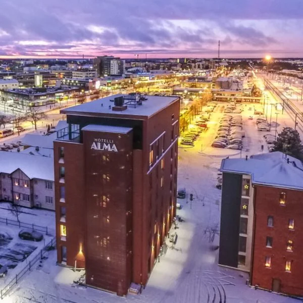 Night view of a snowy cityscape with a hotel named Hotel Alma in the foreground, illuminated streets, and a cloudy purple sky.
