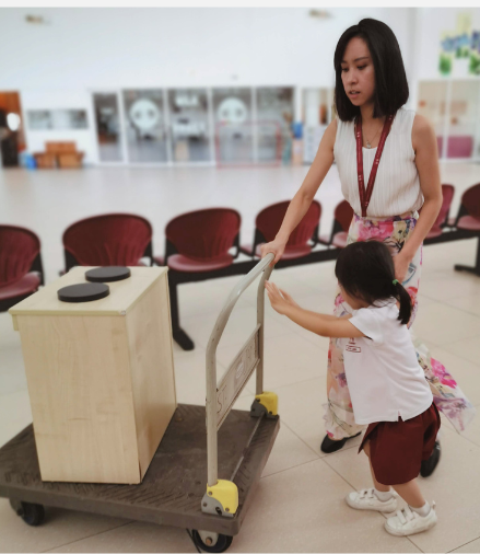 Hermes Lu guiding a young child as they help push a cart carrying furniture.