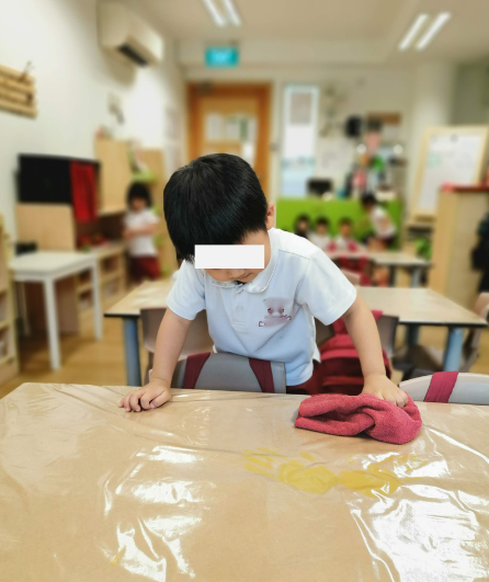 Young child helping to clean a classroom table with a cloth.