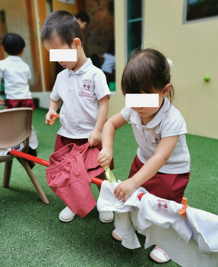 Two toddlers practising fine motor skills by attaching clothes with pegs to a bamboo pole.