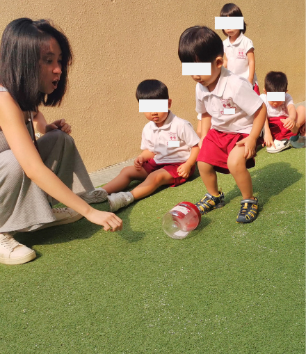 Toddler rolling a plastic container down a small slope while Hermes Lu encourages from nearby.