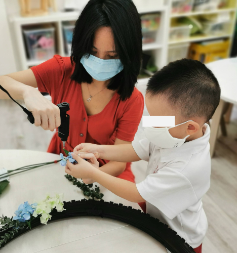 Young child helping hold materials while Hermes Lu applies hot glue during a collaborative project.