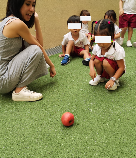 Toddler experimenting with rolling a toy apple down a slope during an outdoor learning activity while Hermes Lu encourages nearby.