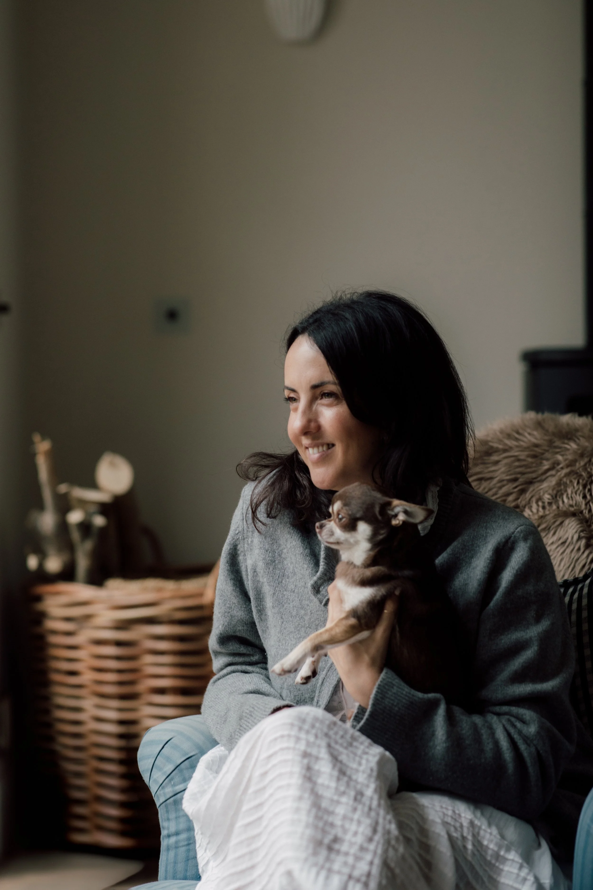 A woman with dark hair smiling and holding a small brown and white dog in a cozy indoor setting.