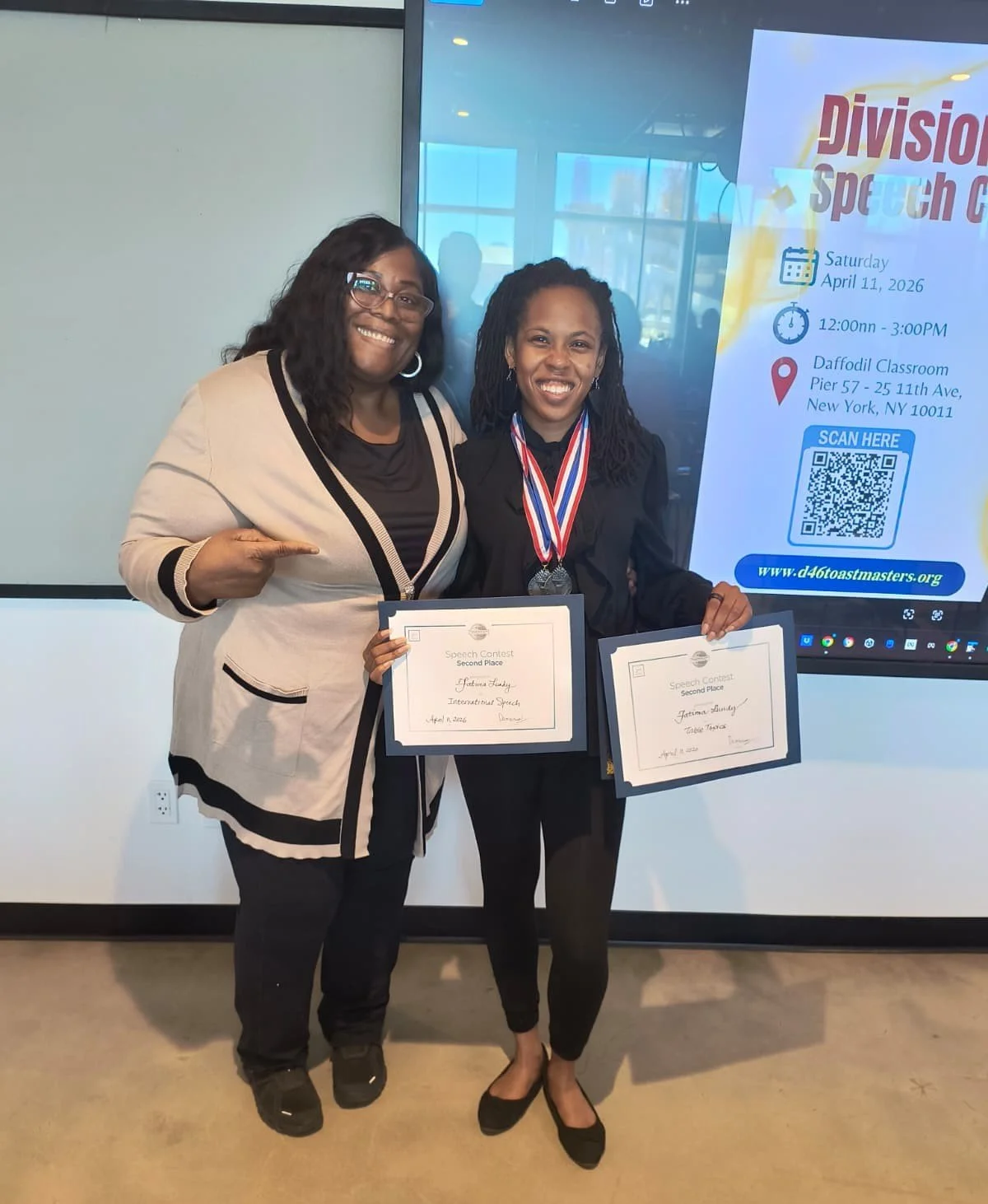 Two women standing together smiling, holding certificates and a medal, at a speech contest award ceremony.
