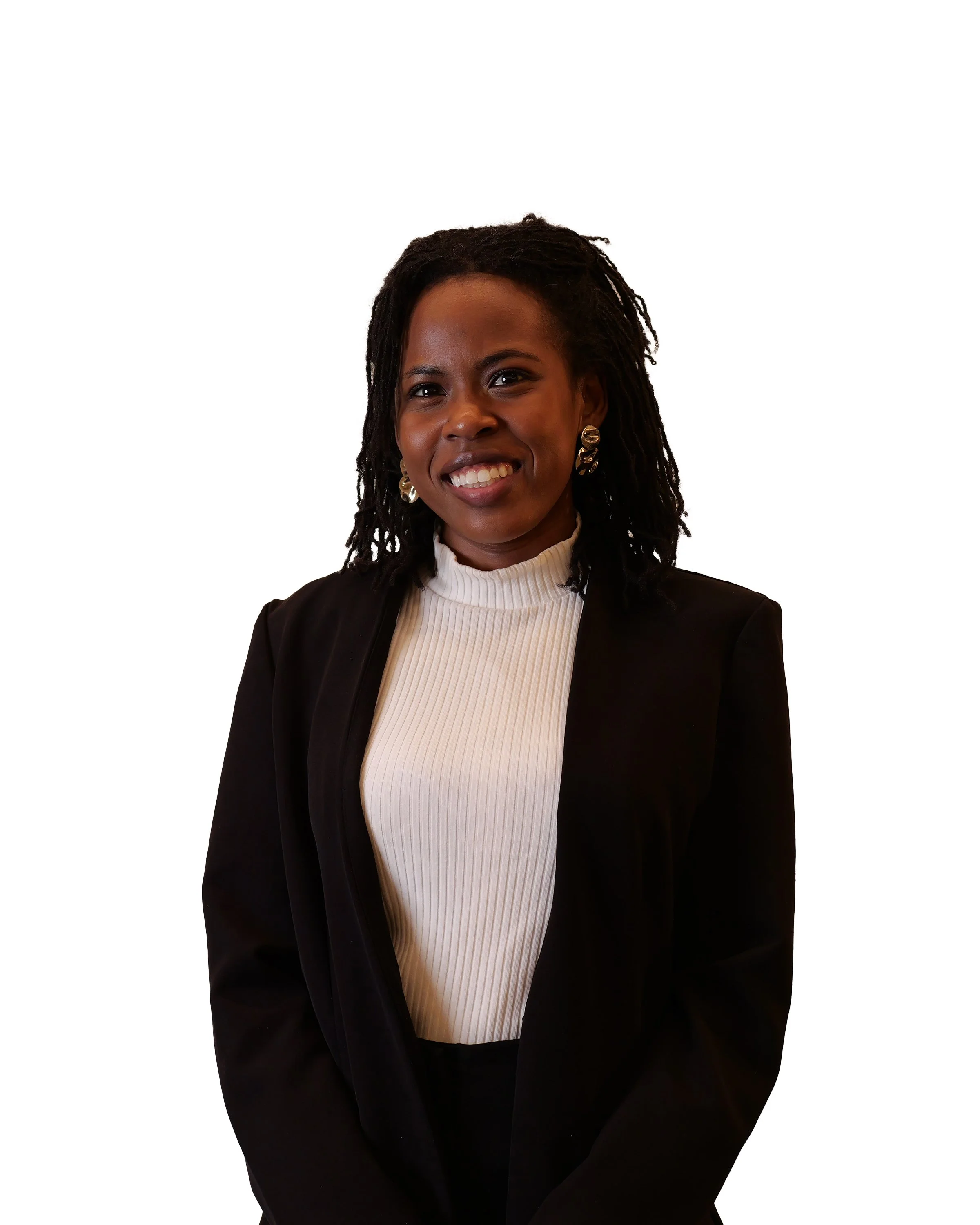 A woman with dark skin and shoulder-length dreadlocks smiling at the camera, wearing a black blazer and a white ribbed turtleneck against a plain white background.