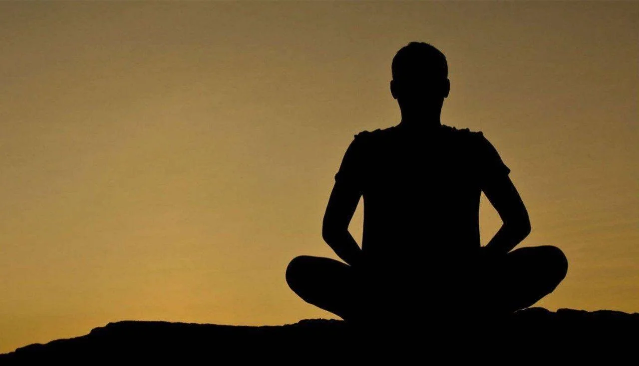 Silhouette of a person sitting in a meditative pose against a sunset or sunrise sky.