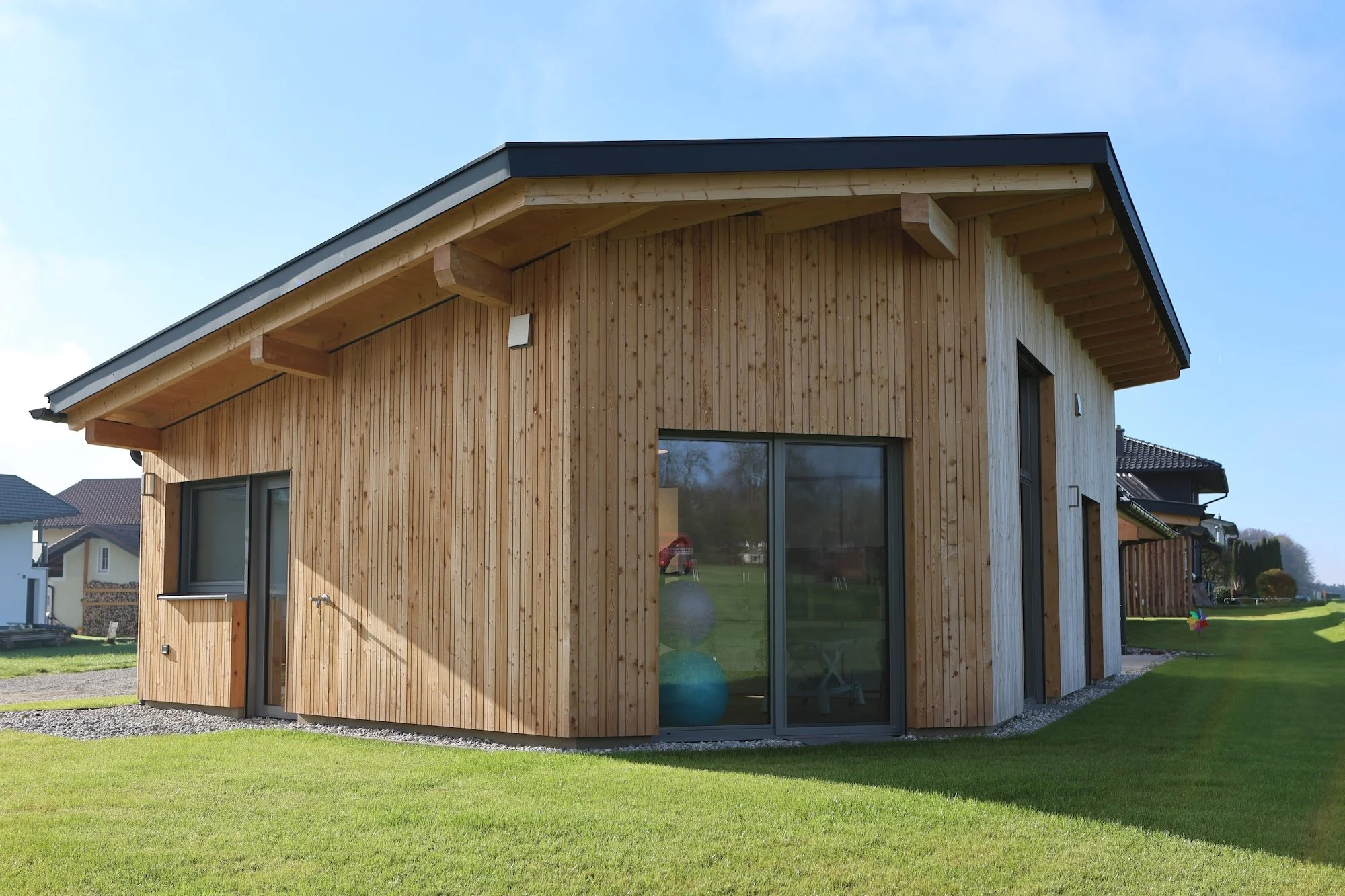Modernes Holzhaus mit großen Fenstern in einem gepflegten Garten bei sonnigem Himmel.