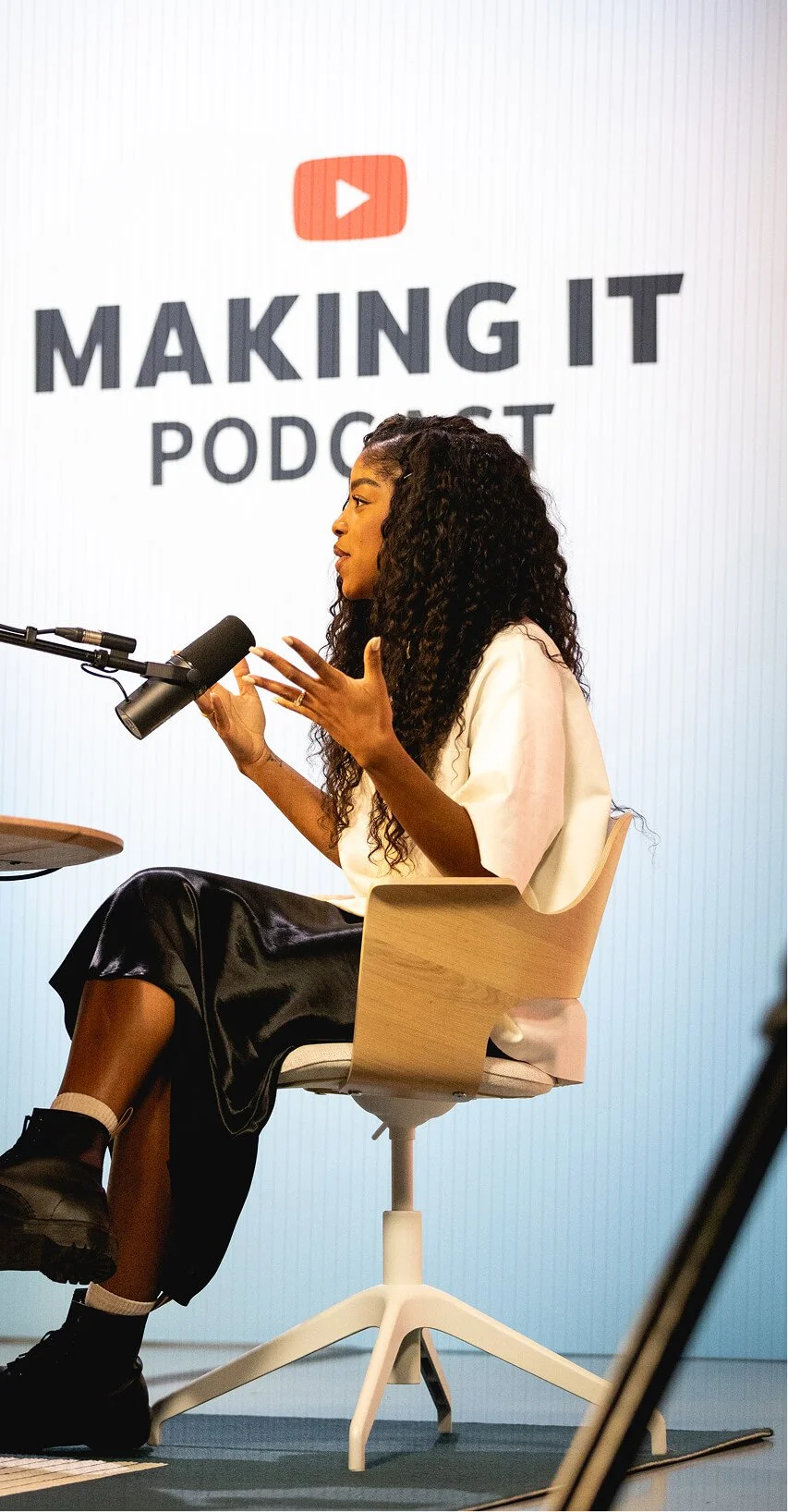 Woman speaking into a microphone during a podcast recording, with a large screen behind her displaying 'YouTube Making It Podcast' logo.