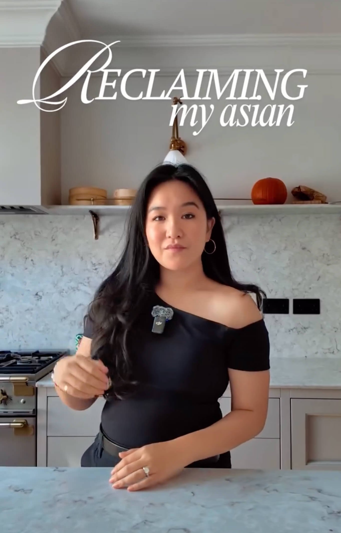 A woman standing in a modern kitchen with a marble countertop, black off-shoulder top, and hoop earrings. There is a pumpkin and a brown bag on the shelf behind her. The text 'Reclaiming my Asian' is written at the top left of the image.