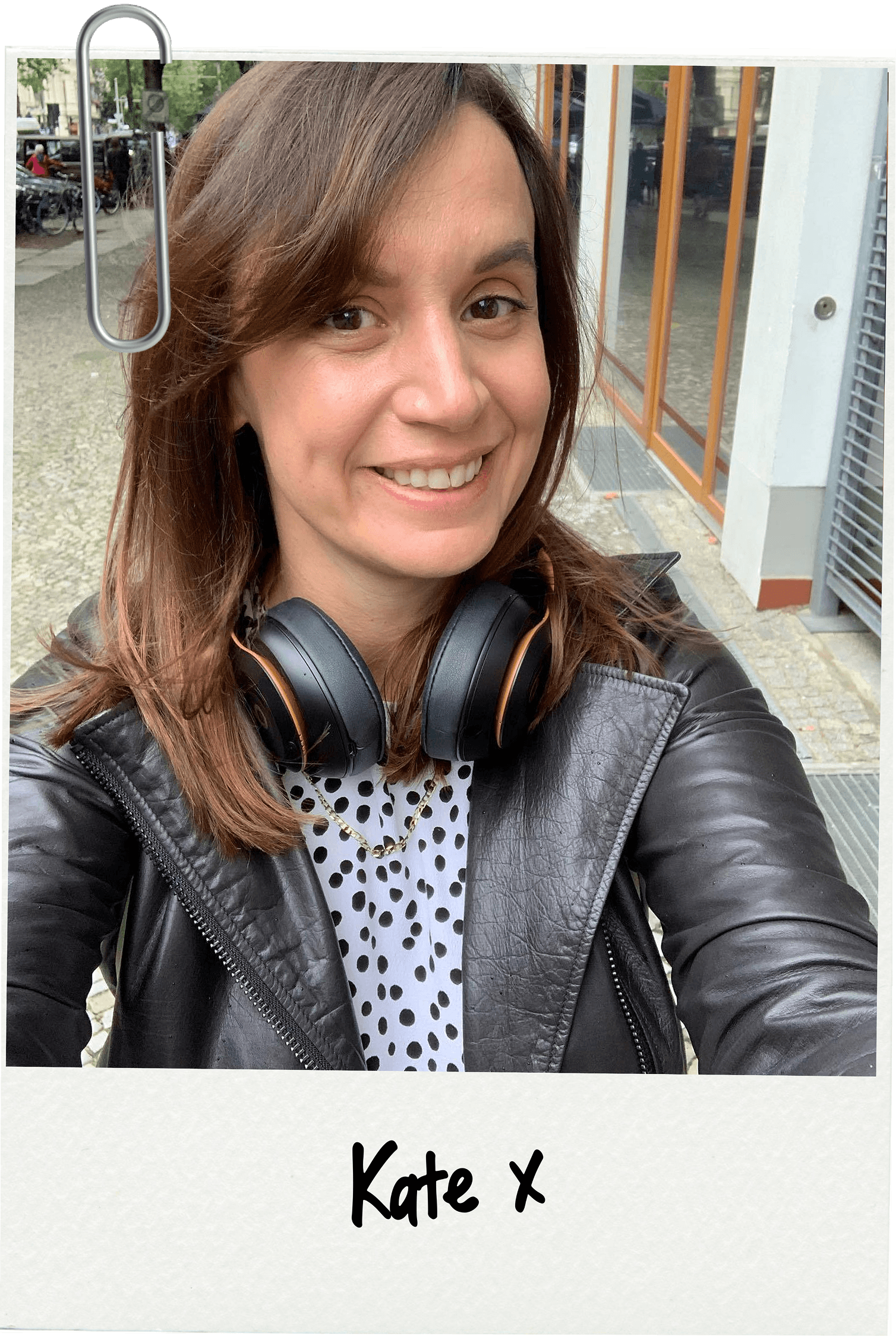 A smiling woman with brown hair, wearing a black leather jacket, polka dot shirt, and headphones around her neck, taking a selfie outdoors on a sidewalk with a storefront and parked bikes in the background.