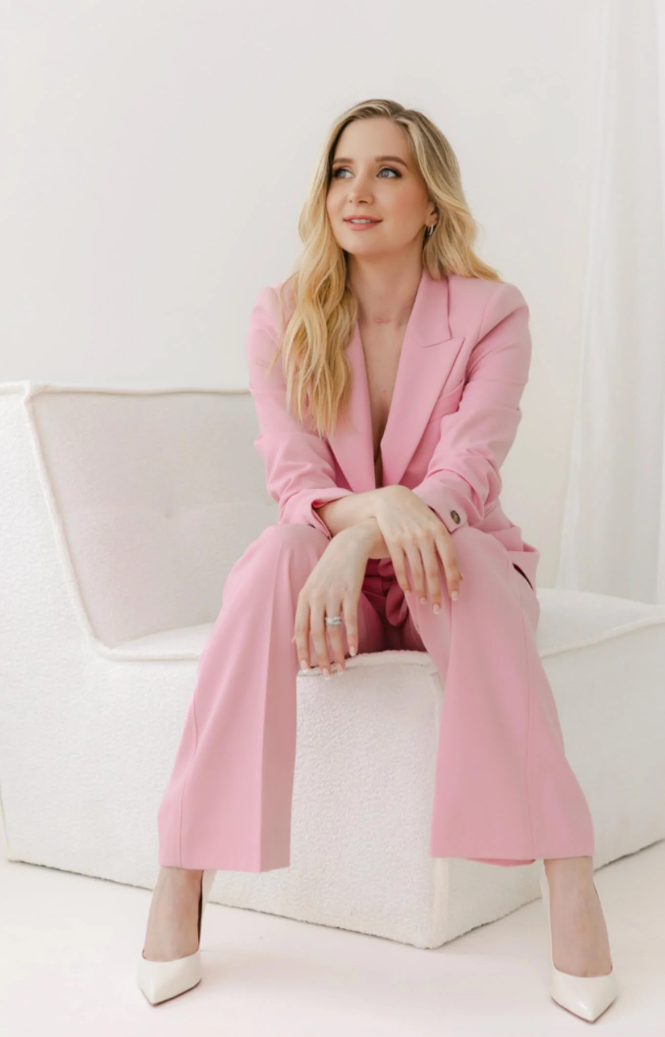 A woman with blonde hair wearing a pink suit and white heels, sitting on a cream sofa, looking to her right with a slight smile.