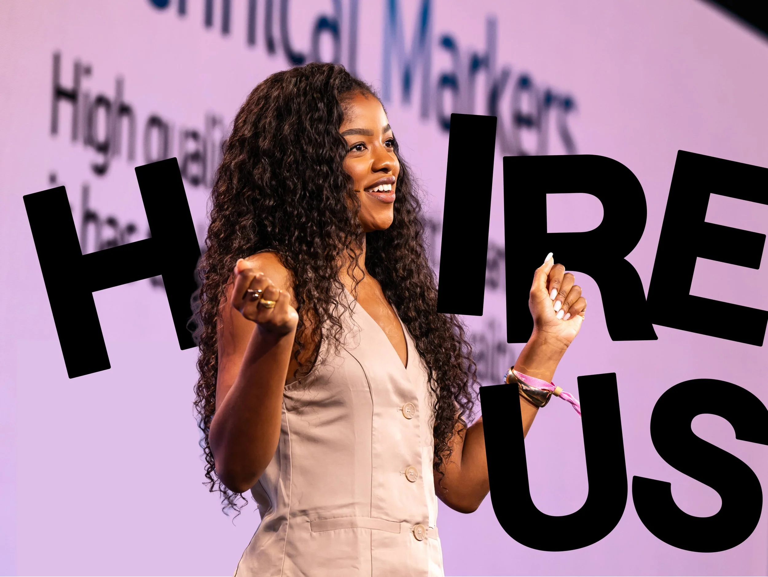 A smiling woman with long curly hair on stage, holding up her fists as if in celebration, in front of a purple background with large black text that says 'HIRE US'.