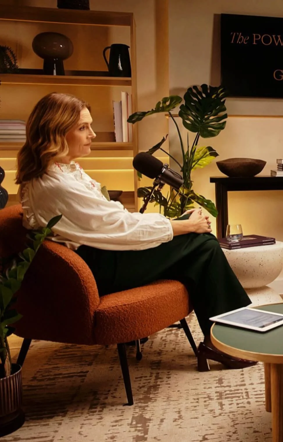 A woman with wavy, shoulder-length hair sits on an orange chair in a cozy room with modern decor, holding a microphone. There are plants, books, a glass of water, and a tablet on a nearby table, with a screen on the wall behind her.