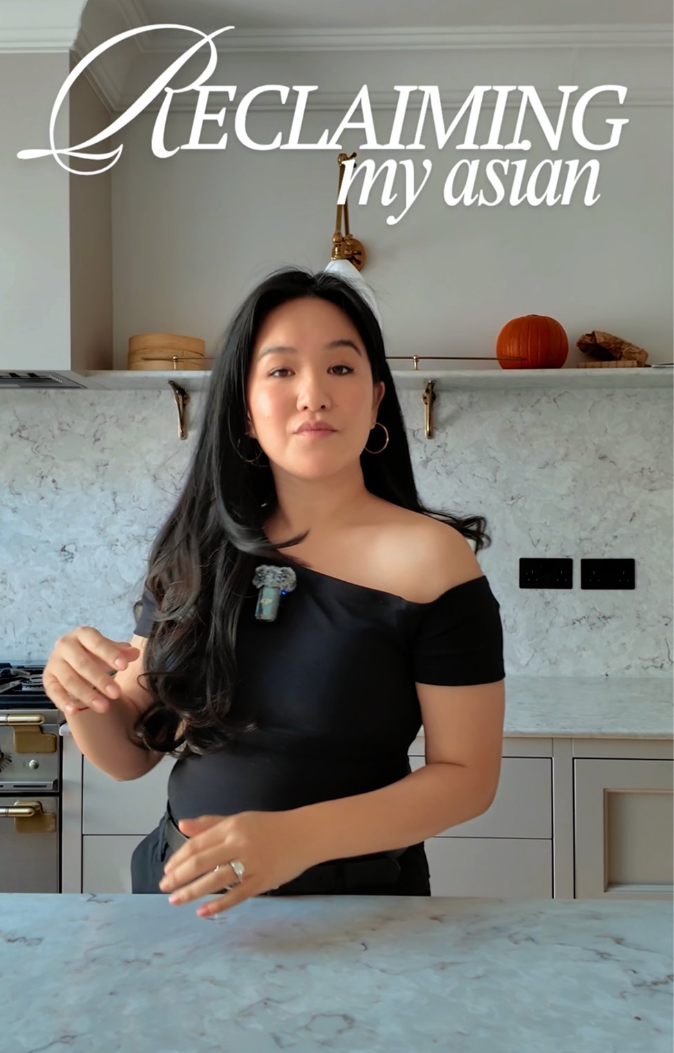 A woman with long black hair, wearing an off-shoulder black top, standing in a modern kitchen with a marble countertop. The background includes a white backsplash, decorative items like a pumpkin and a basket on a shelf, and a chandelier. Text overlay reads, "Reclaiming my Asian."