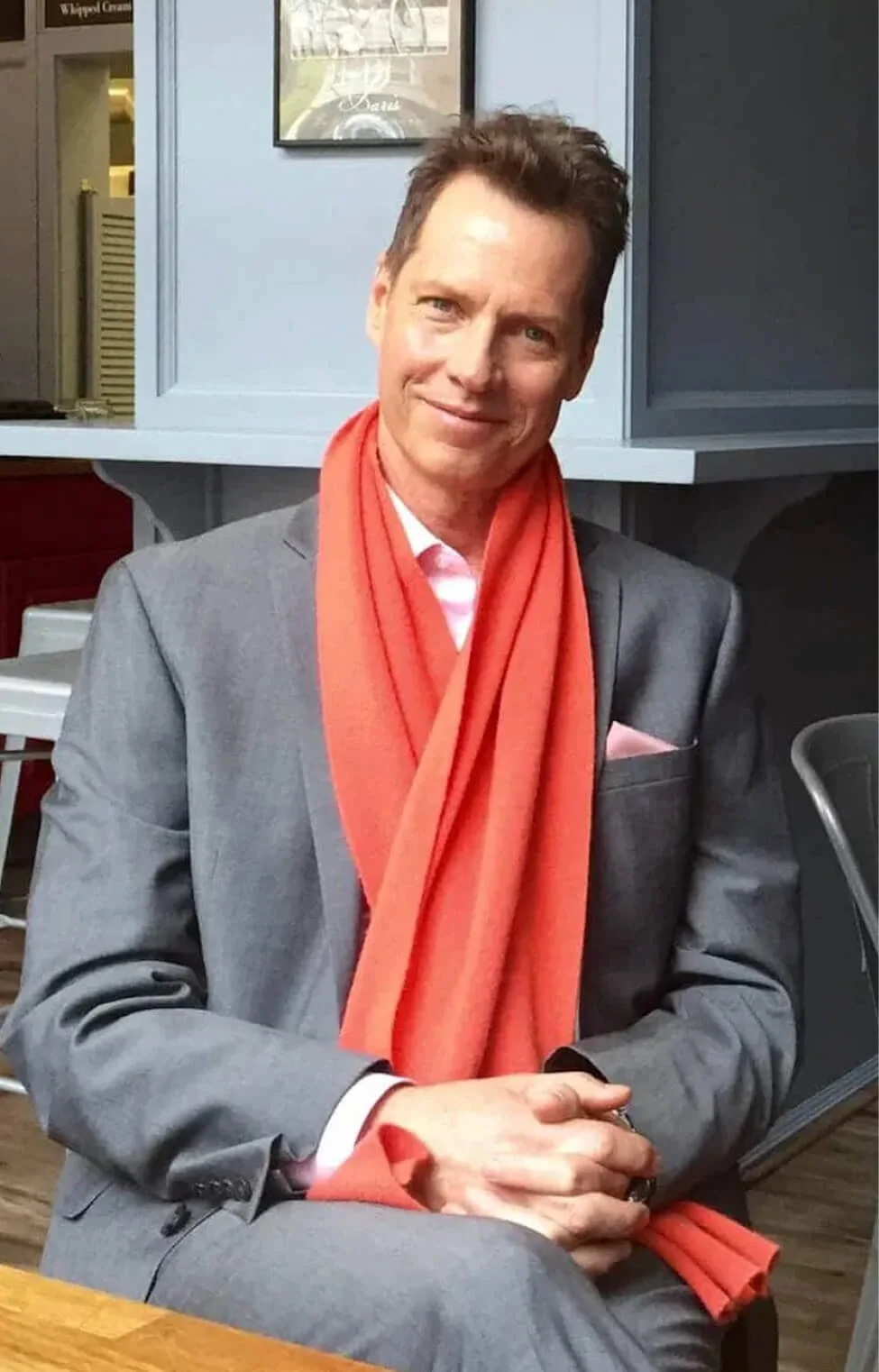 A man in a gray suit with a pink shirt and pink pocket square, wearing an orange scarf, sitting at a table in a cafe or restaurant.