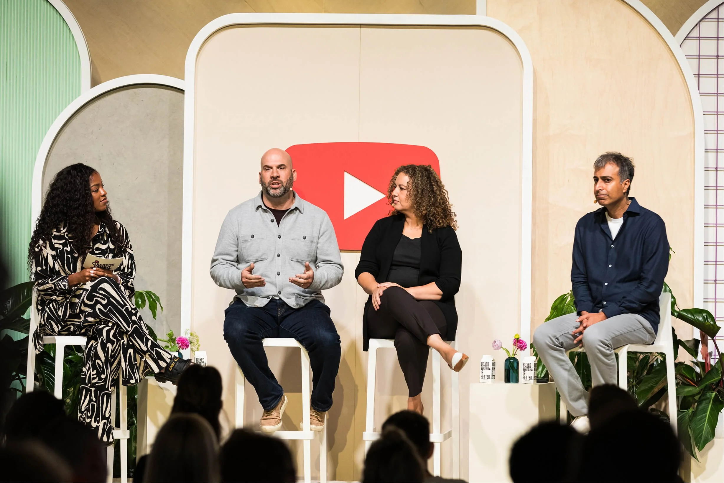 Panel discussion with four diverse speakers on stage, seated on white chairs, with a backdrop featuring a large YouTube play button logo. Audience members are visible in the foreground.