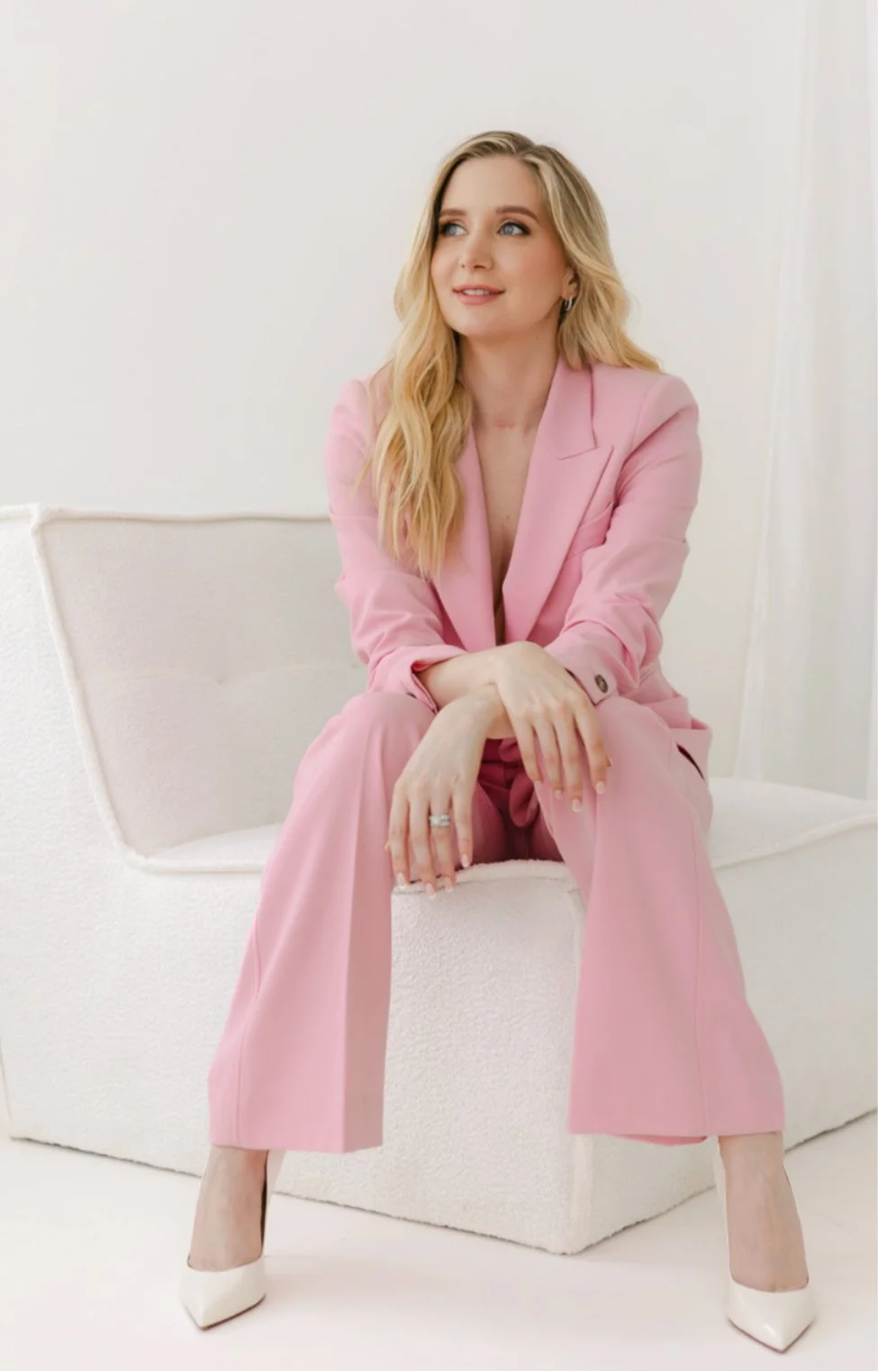 A woman with blonde hair sitting on a white sofa, wearing a pink suit and white heels, smiling softly and looking to her left.