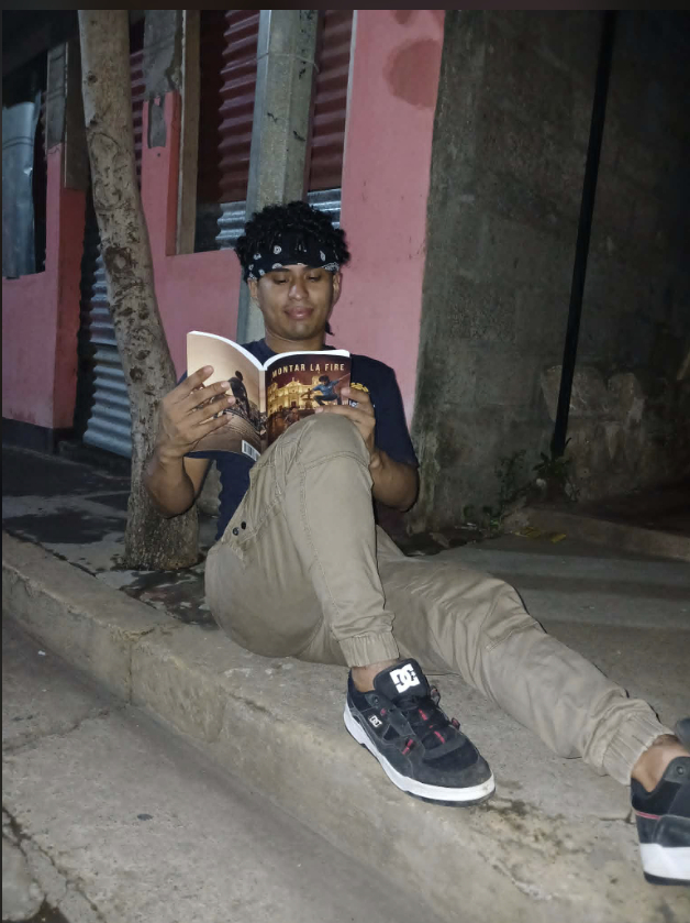 A young man with curly hair and a bandana reading a book while sitting on the sidewalk at night, near a tree and a pink building with corrugated metal siding.