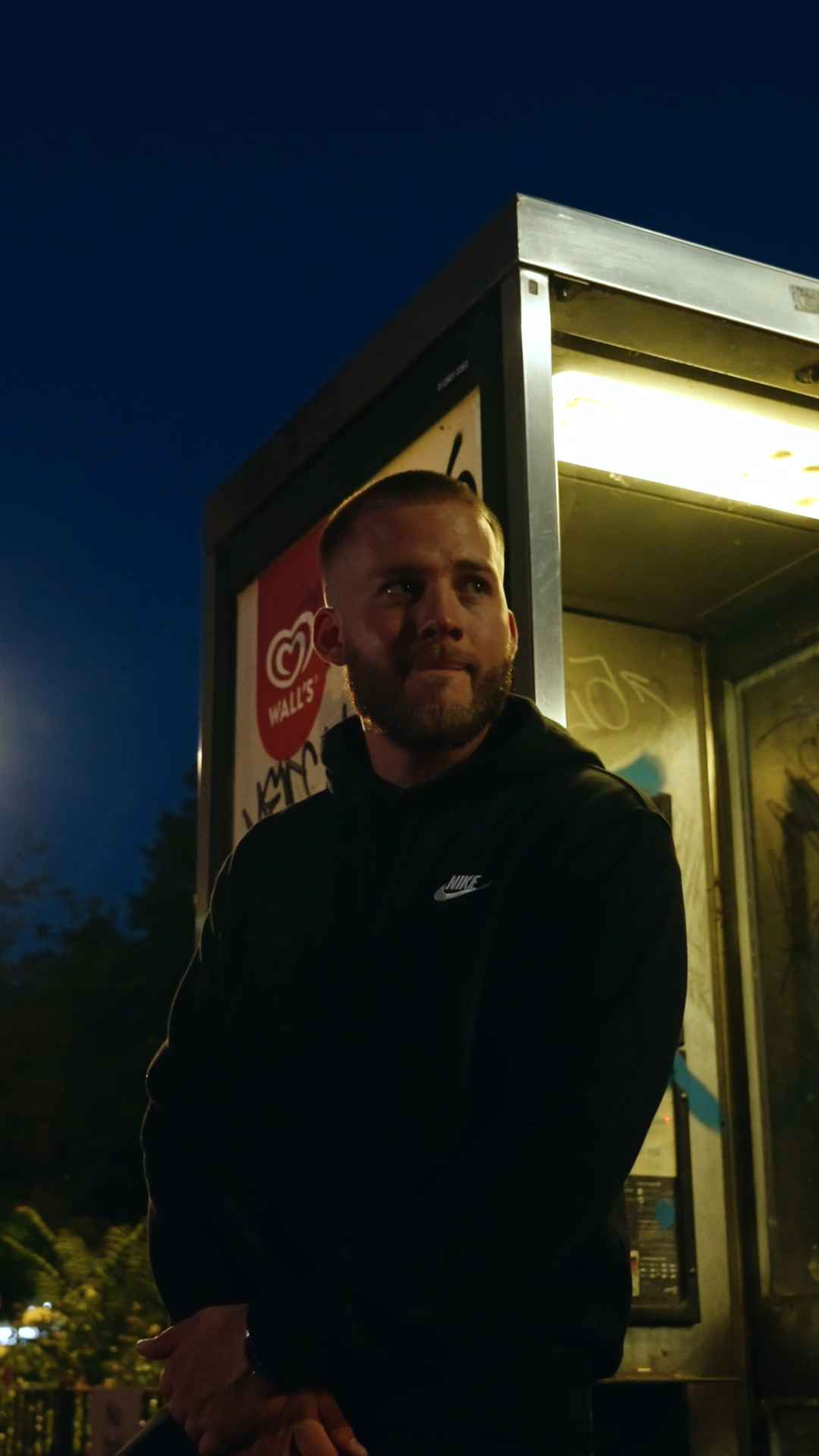 A man with a beard in a black Nike jacket standing outside near a lit kiosk or phone booth at night, with a dark blue sky in the background.