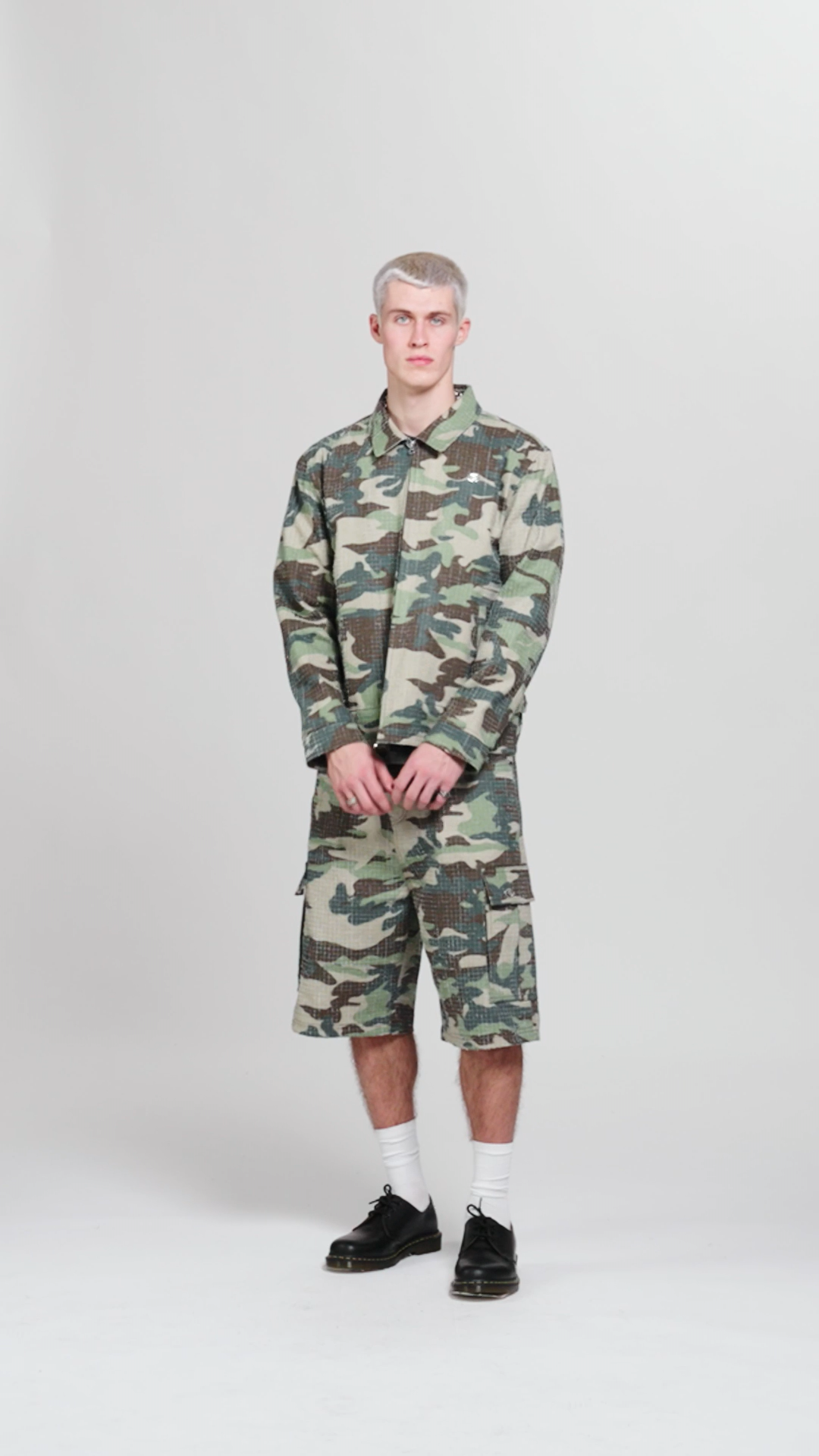 A young man with short blond hair stands against a plain white background, wearing a green and brown camouflage jacket and matching shorts, white socks, and black shoes.