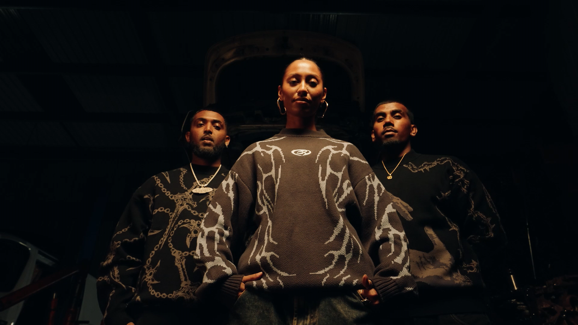 Three people standing below a vehicle engine in a dark garage, the woman in the center is wearing a patterned sweater, and the two men on either side are wearing black shirts with gold jewelry.