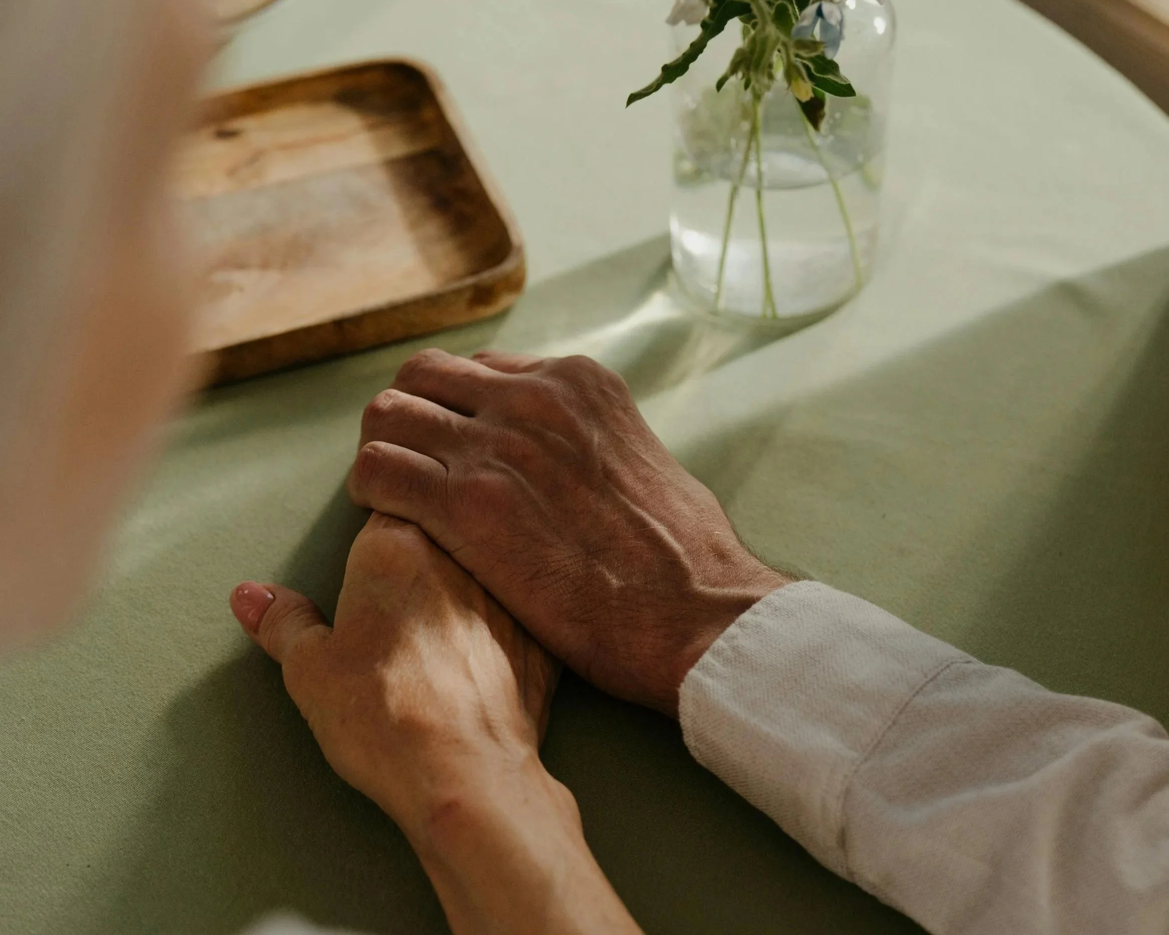Close-up view of two elderly hands gently holding each other on a light green surface, with a glass vase containing a small bouquet of flowers in the background.