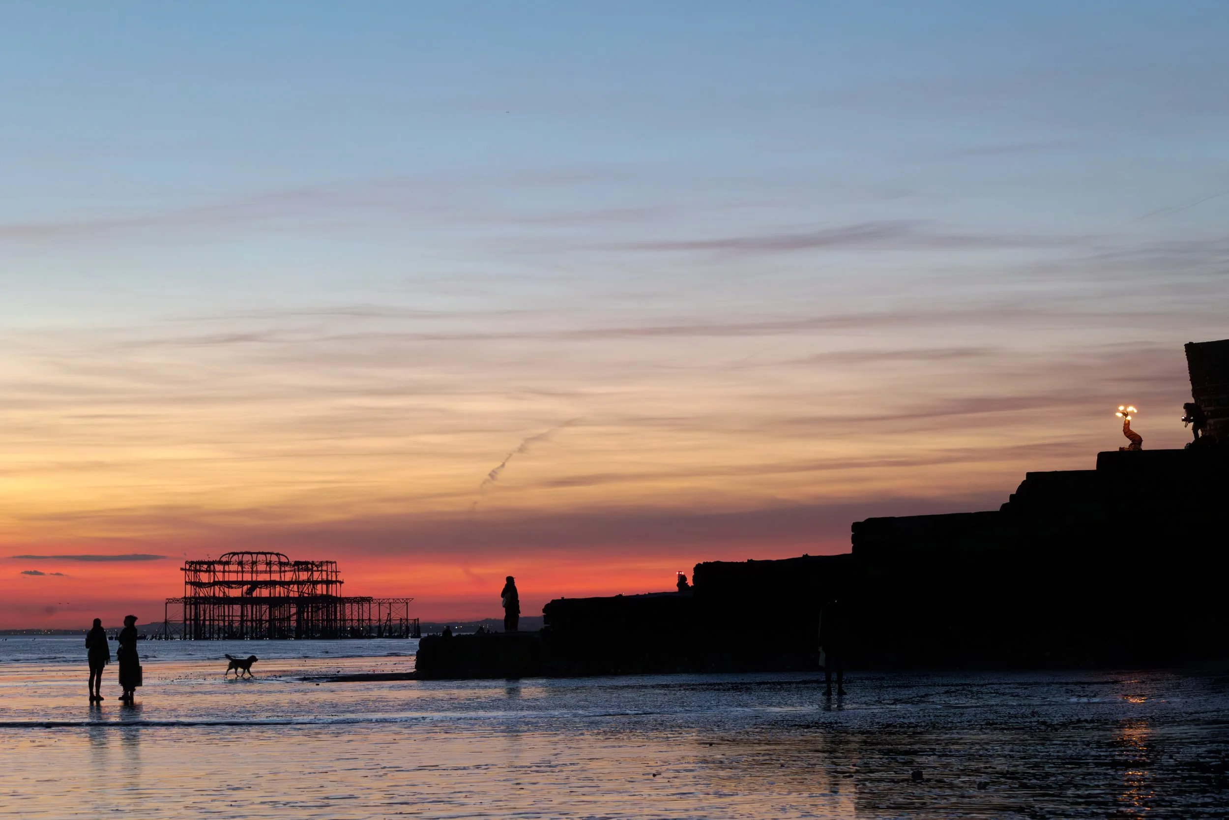 Silhouettes of people and a dog on Brighton beach during sunset with the West Pier structure in the background.