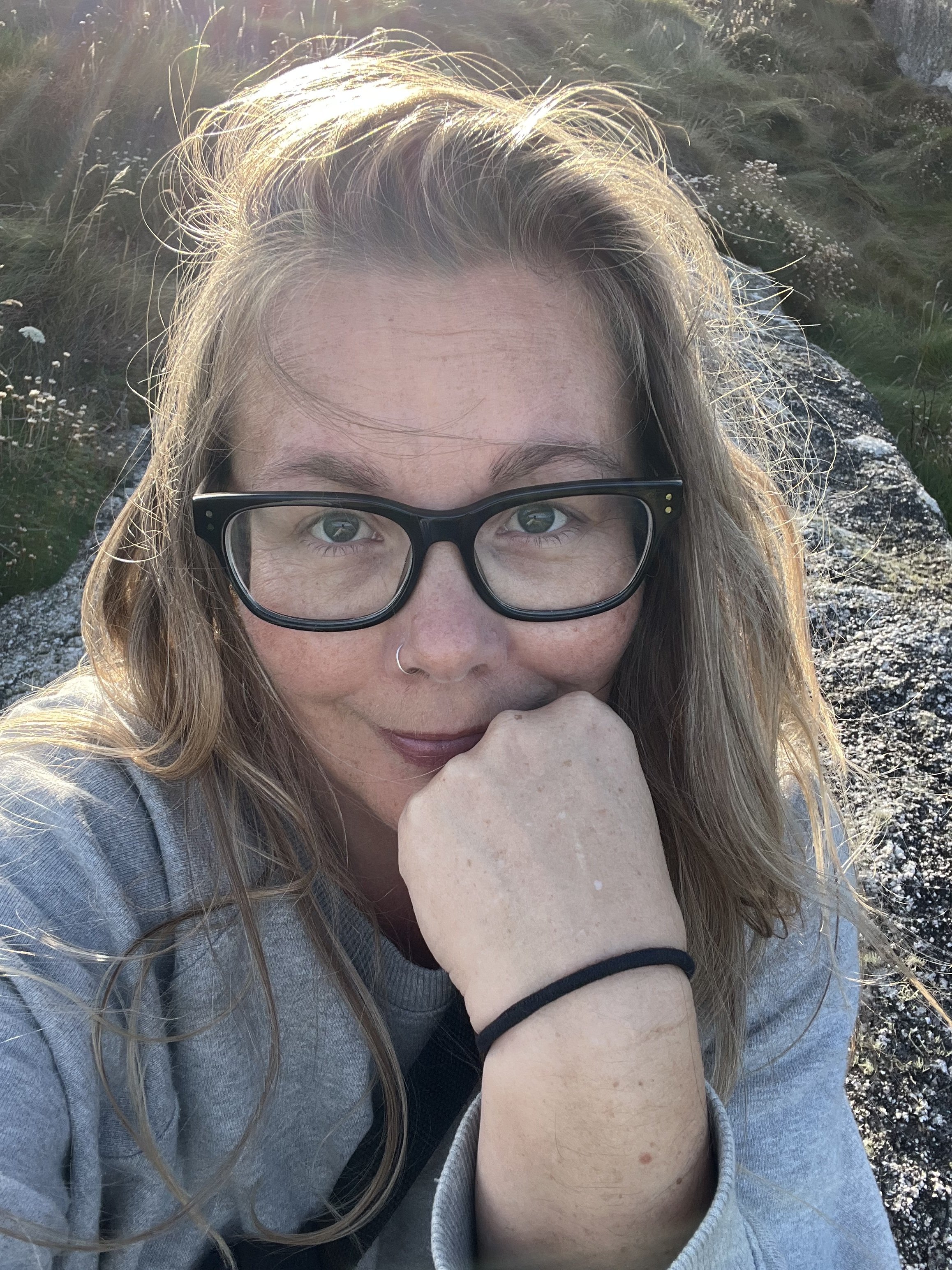 A woman with long wavy hair, black glasses, and a nose ring is resting her chin on her hand outdoors. She is wearing a gray sweatshirt, and the background features rocky terrain and green grass, illuminated by sunlight.