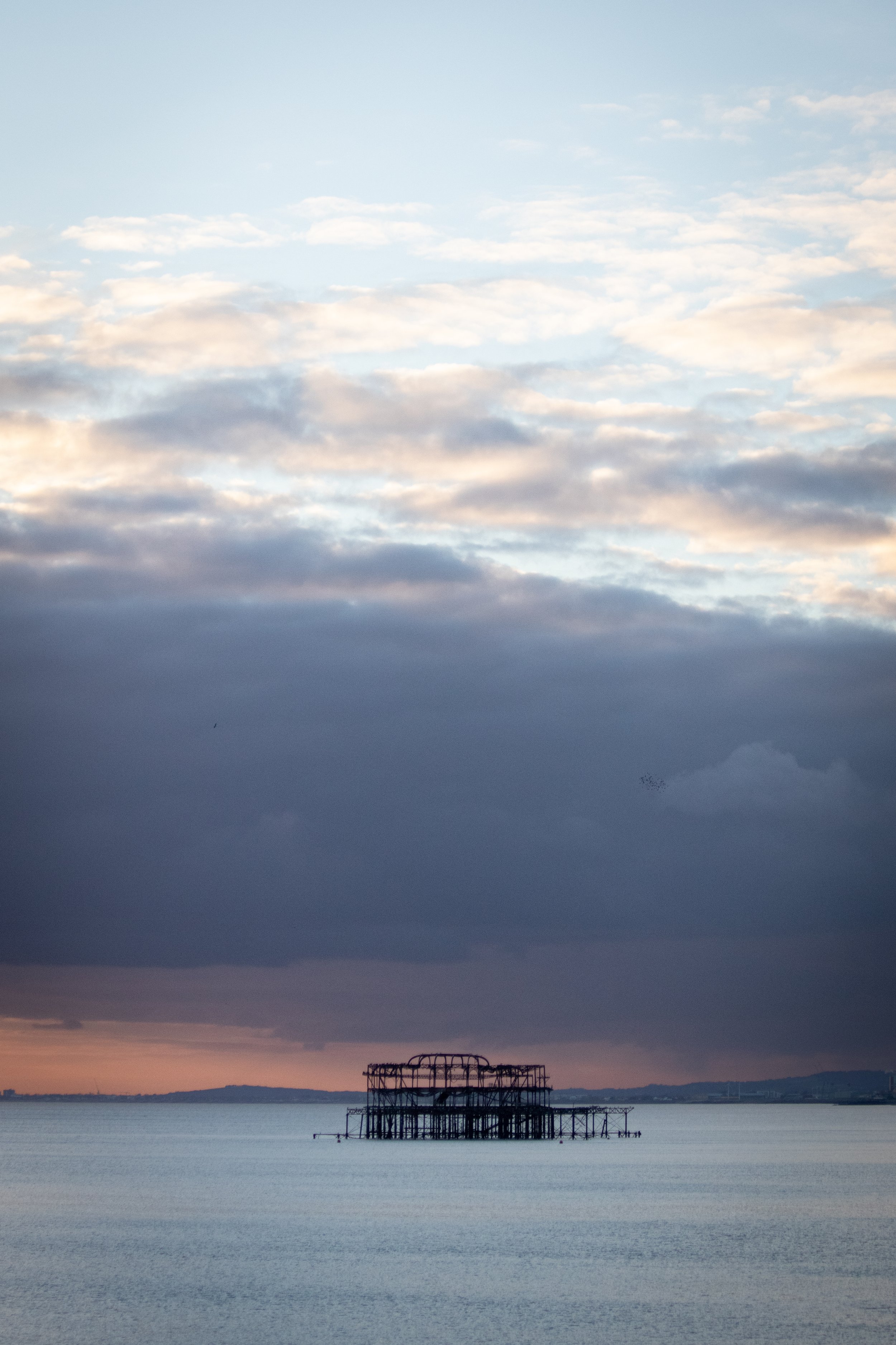 Brighton's West Pier partially submerged in calm water, with a background of a dramatic cloudy sky and distant shoreline under a soft sunset.