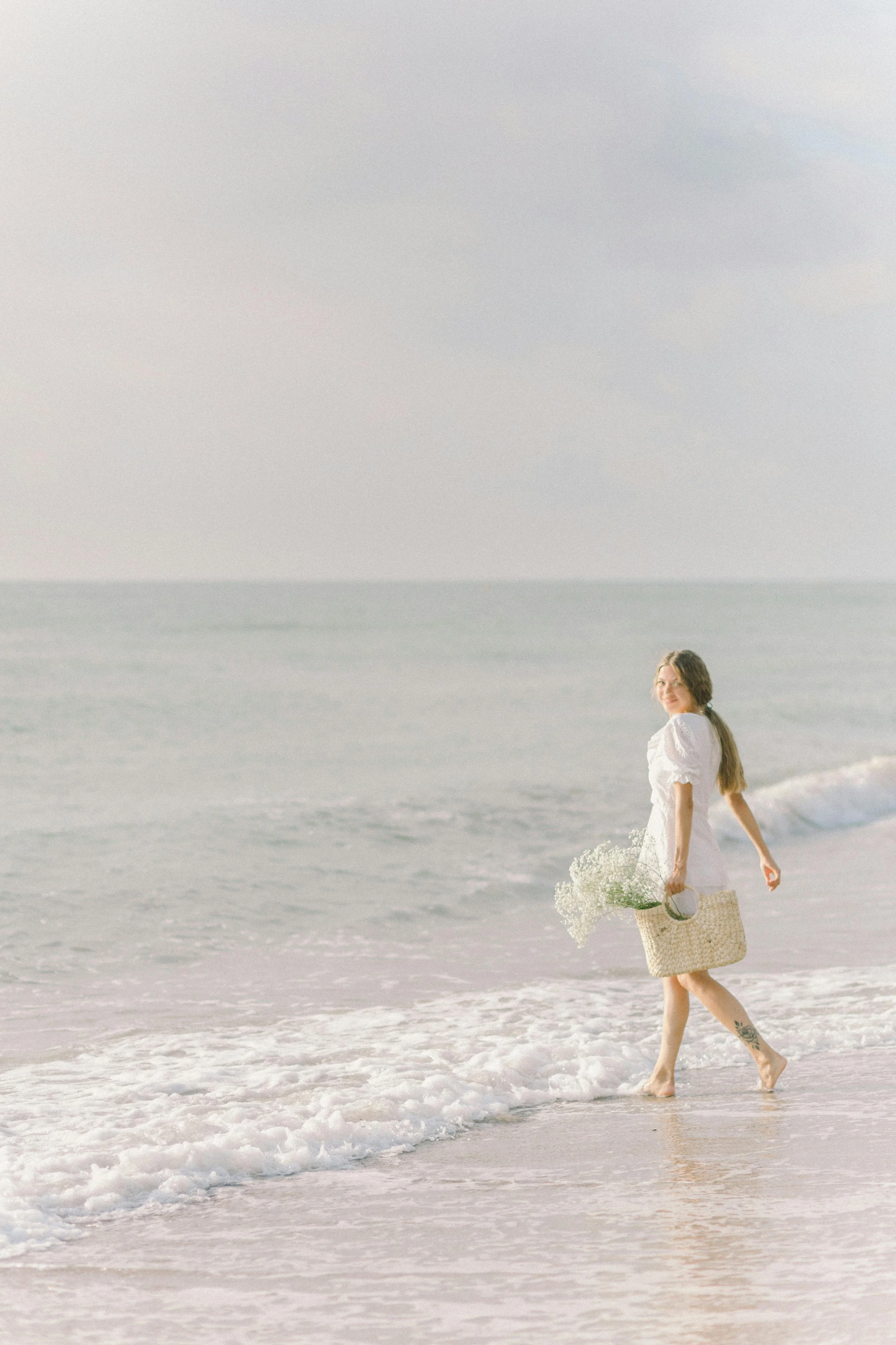 A woman in a white dress walking along the shoreline of the beach with a basket of white flowers.