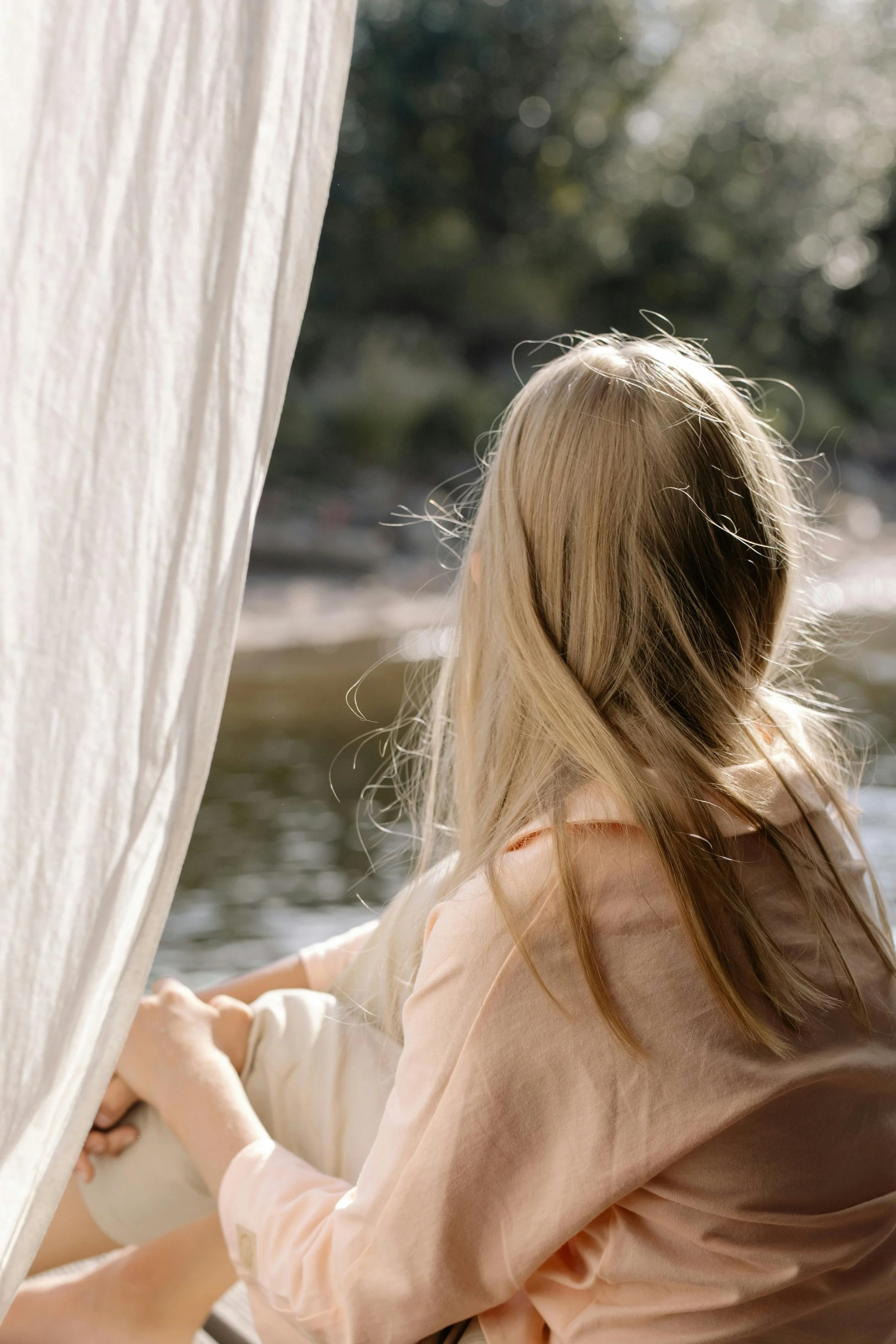 A young girl with blonde hair sitting outside near a body of water, looking away from the camera. She is partially covered by a light-colored fabric curtain.