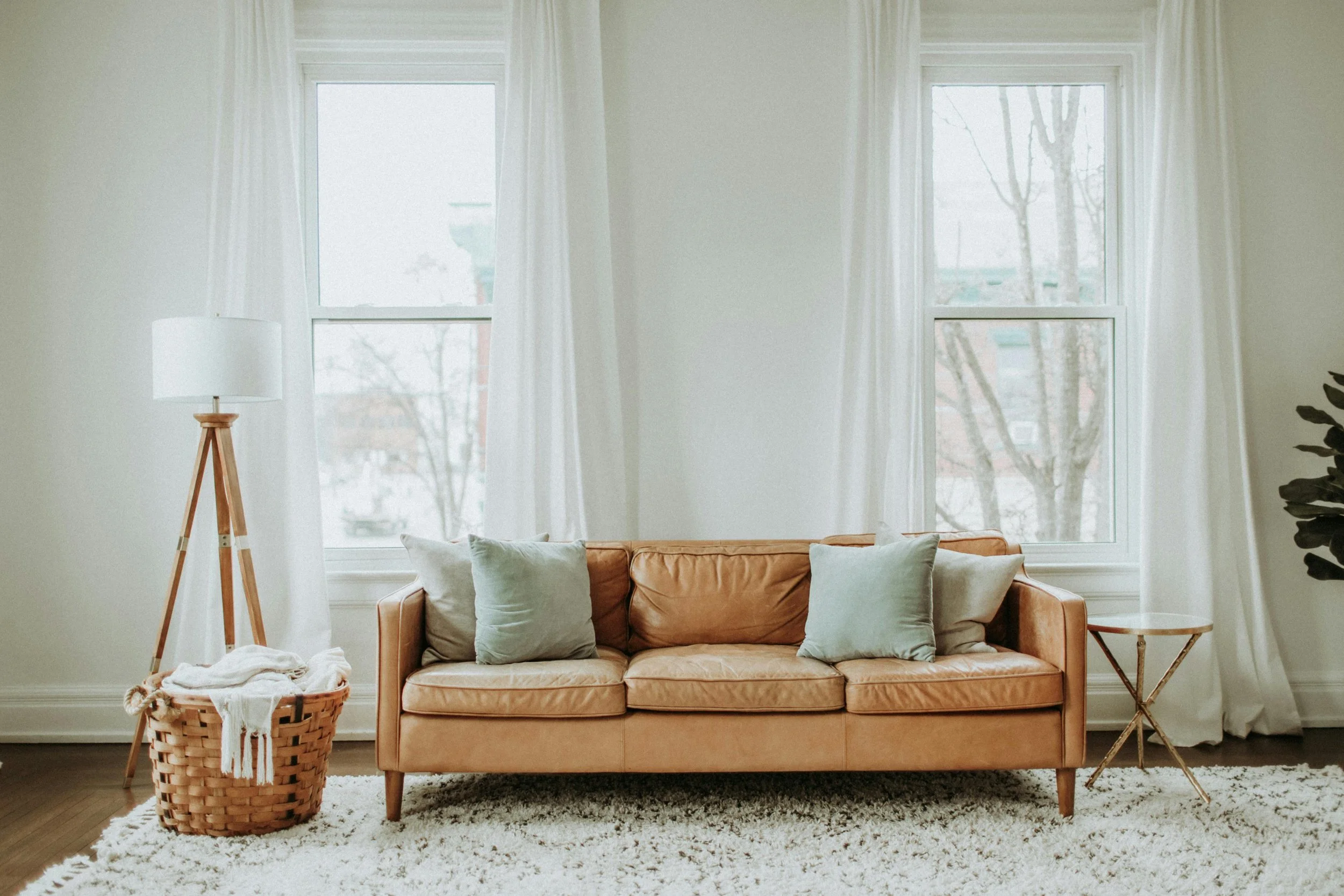 A living room with a tan leather sofa, four throw pillows, two large windows with white curtains, a tall floor lamp with a wooden tripod stand, and a small side table, all on a white textured rug.