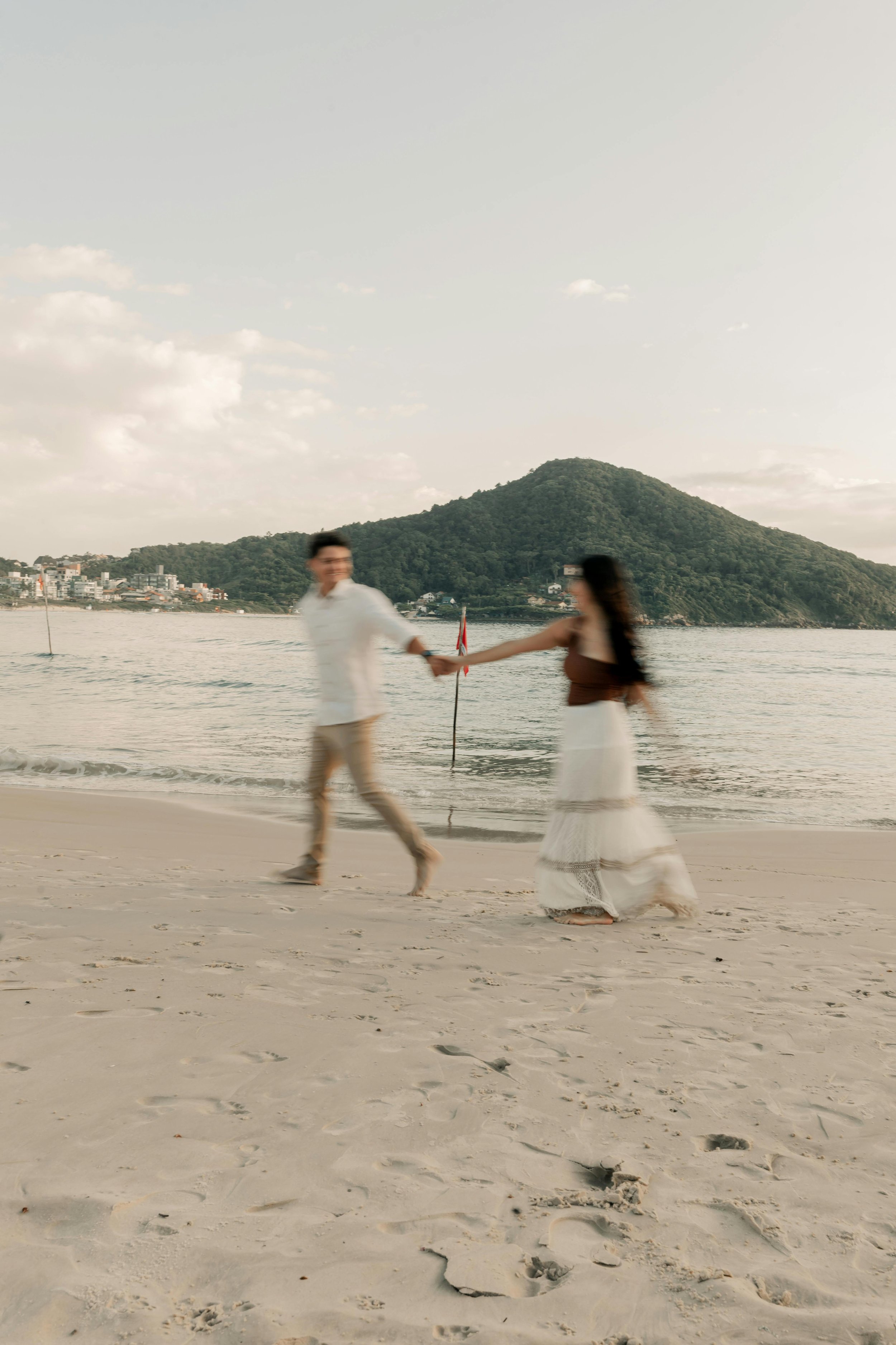 A couple walking on the beach holding hands, with a mountain and water in the background, during the daytime.