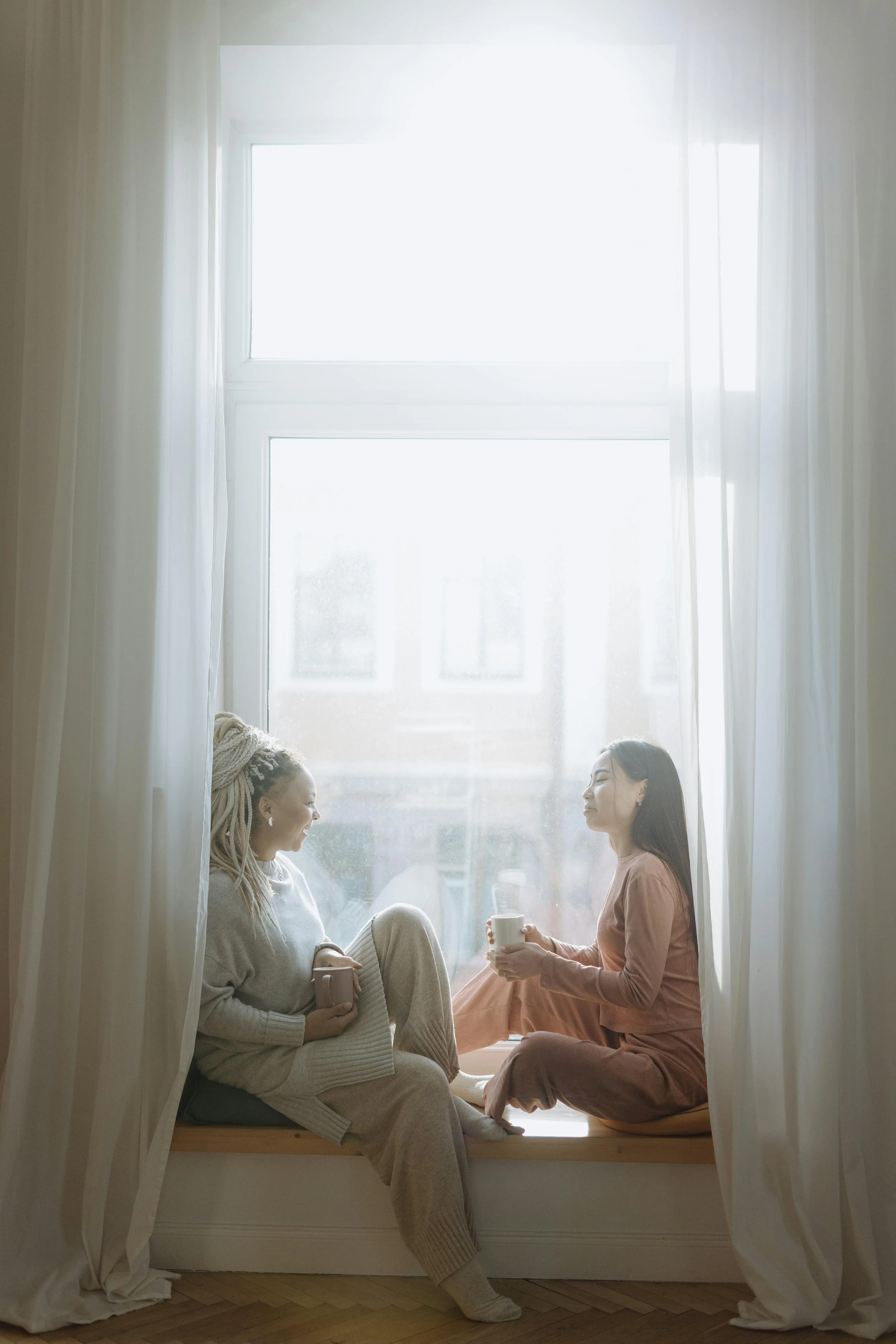 Two women sitting on a cushioned window seat, chatting and holding cups, with a large window and curtains in the background.