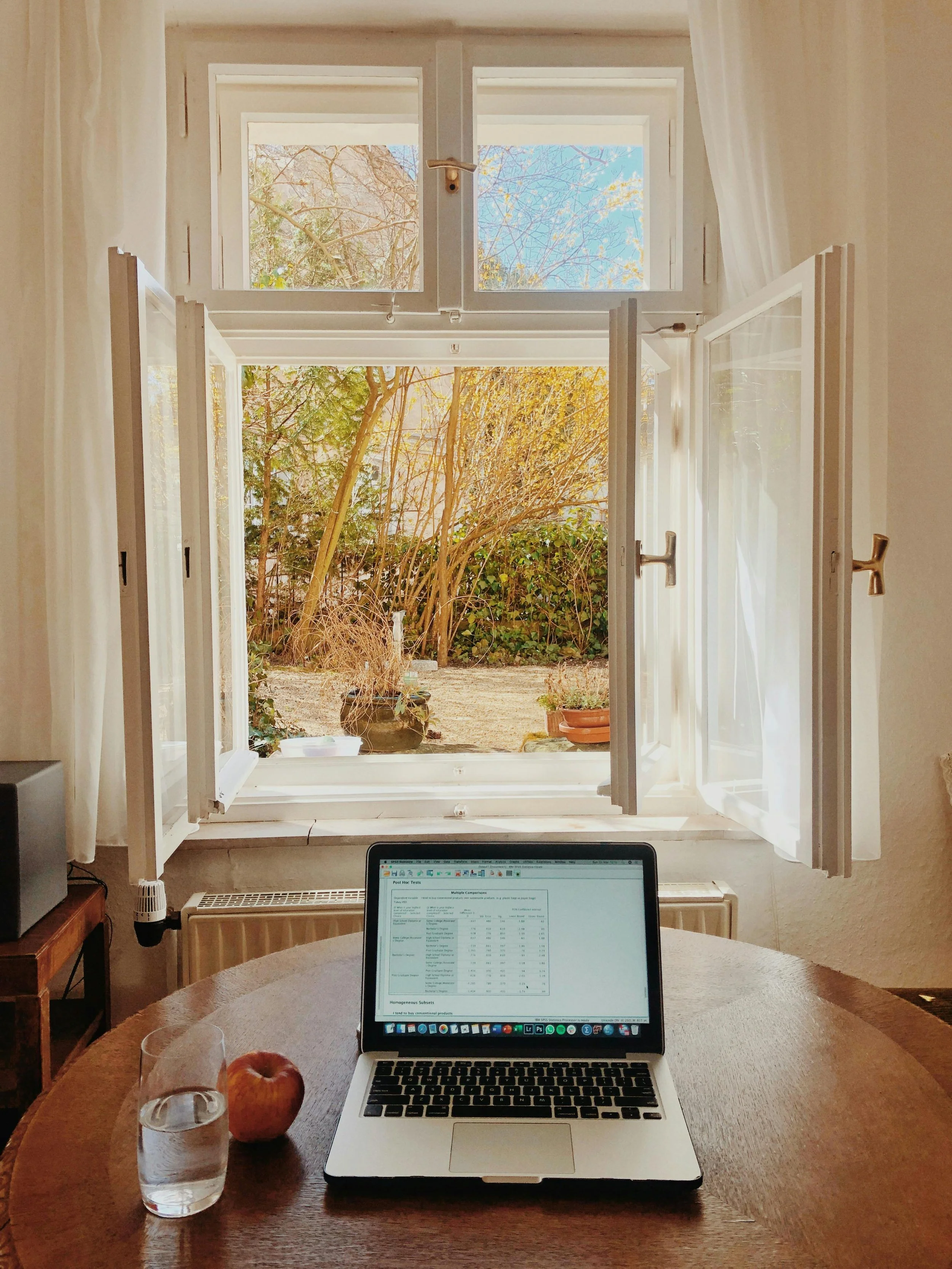 A laptop open on a wooden table with a glass of water and an apple beside it, in front of an open window showing a garden with plants and trees outside.