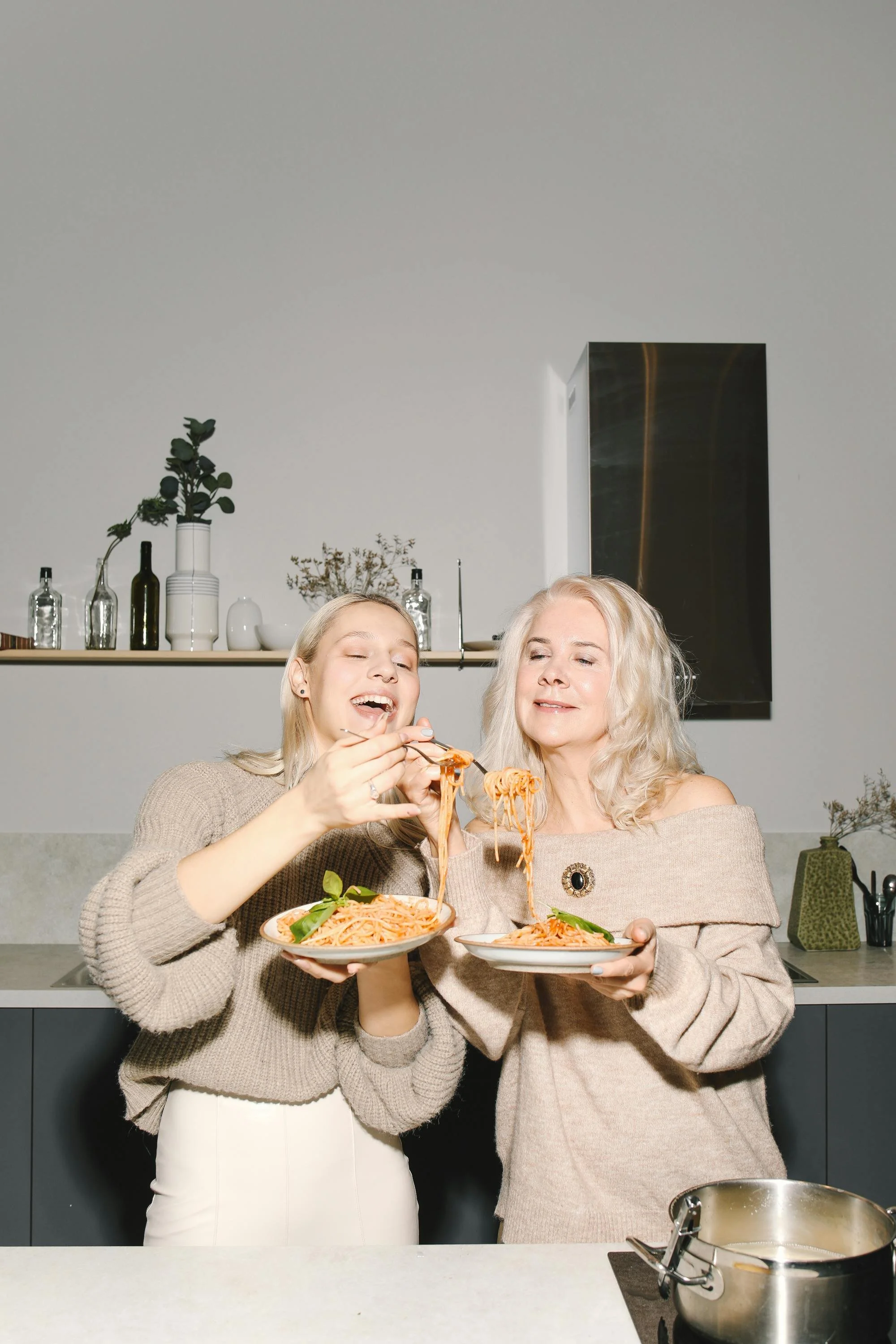 A young woman and an older woman enjoy spaghetti together in a modern kitchen, smiling and sharing a meal.
