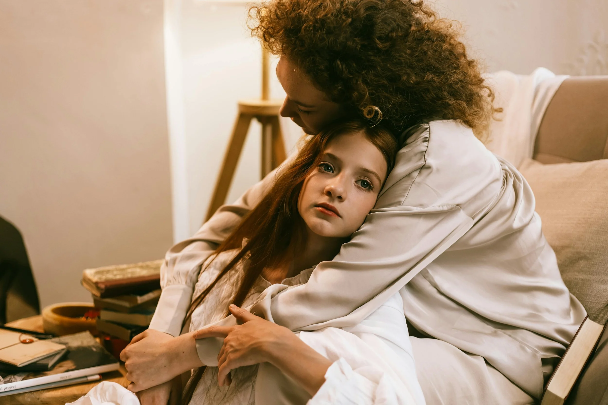 A woman comforting a young girl who appears sad or upset, sitting on a bed with books and a notebook nearby.