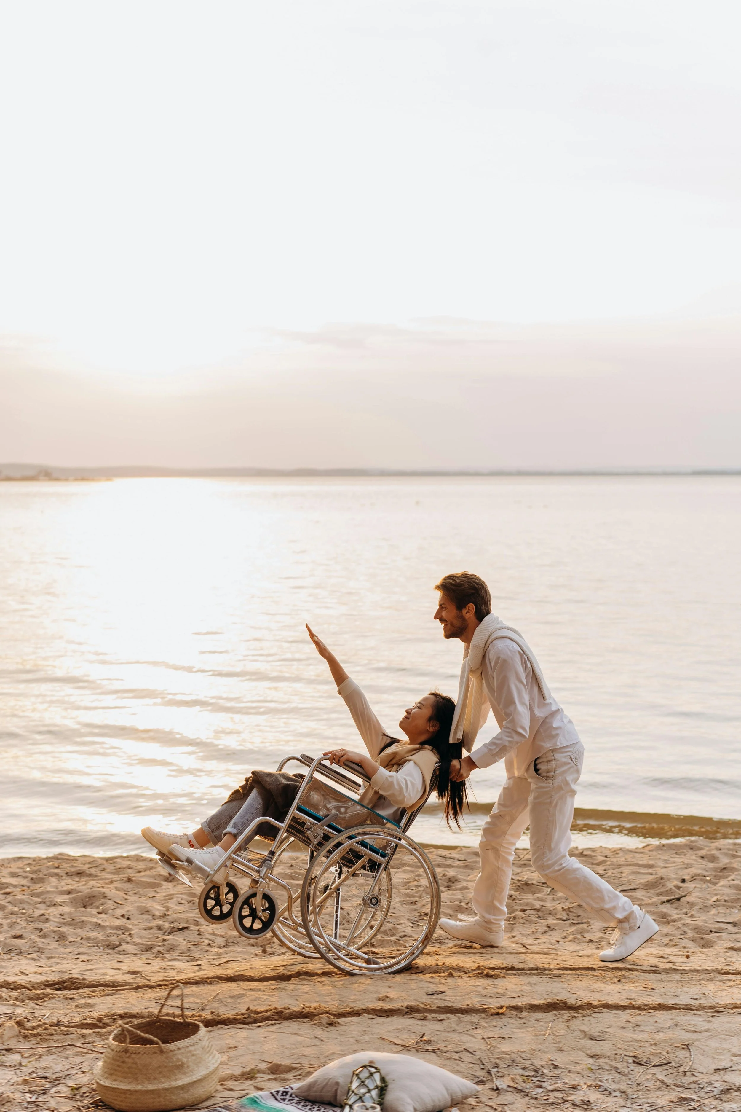 A man and woman, one in a wheelchair, enjoying a moment together on a beach during sunset, with water and sky in the background.