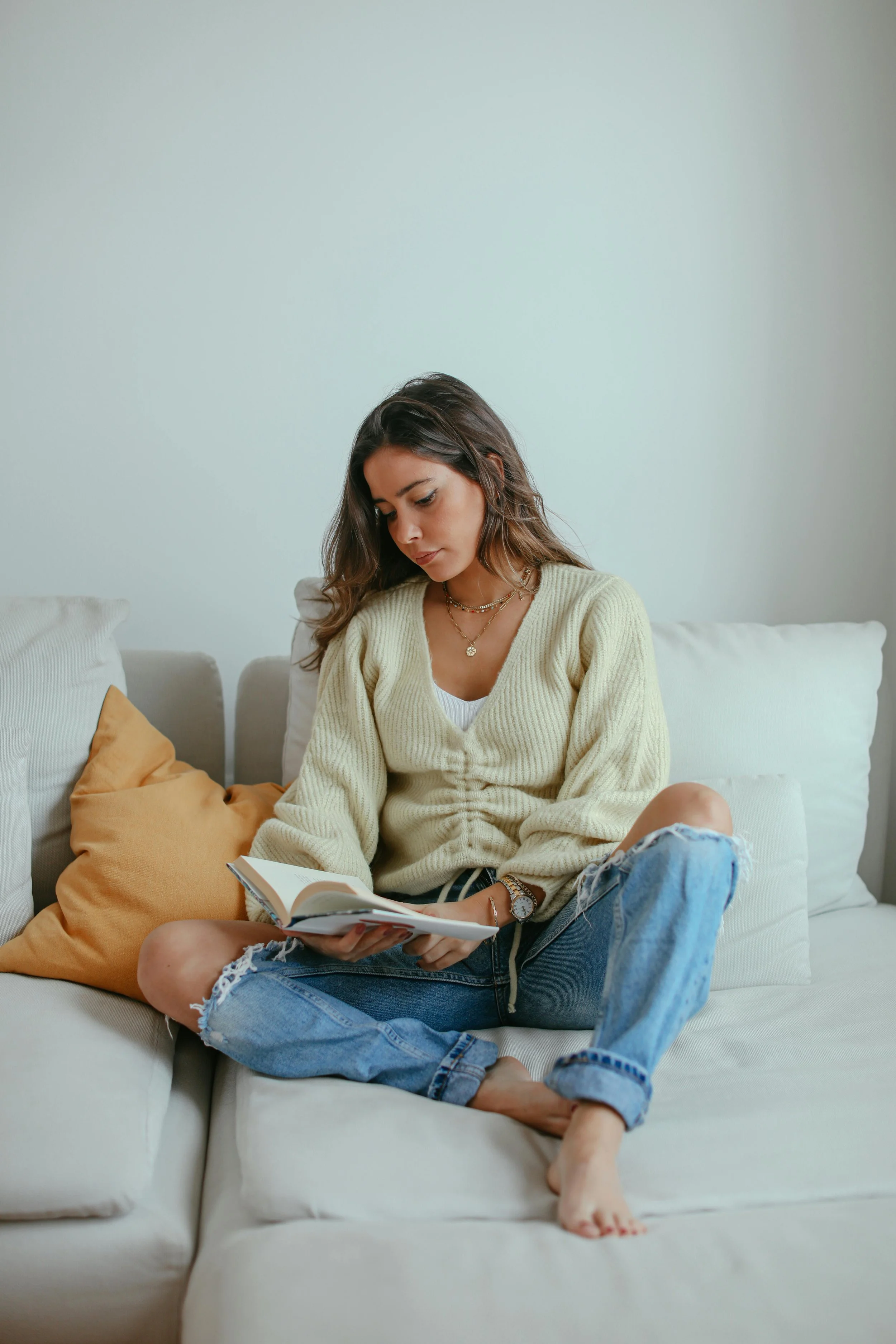 A young woman sitting on a white sofa, reading a book, wearing a cream sweater and ripped jeans, with pillows behind her, in a minimalistic room with white walls.