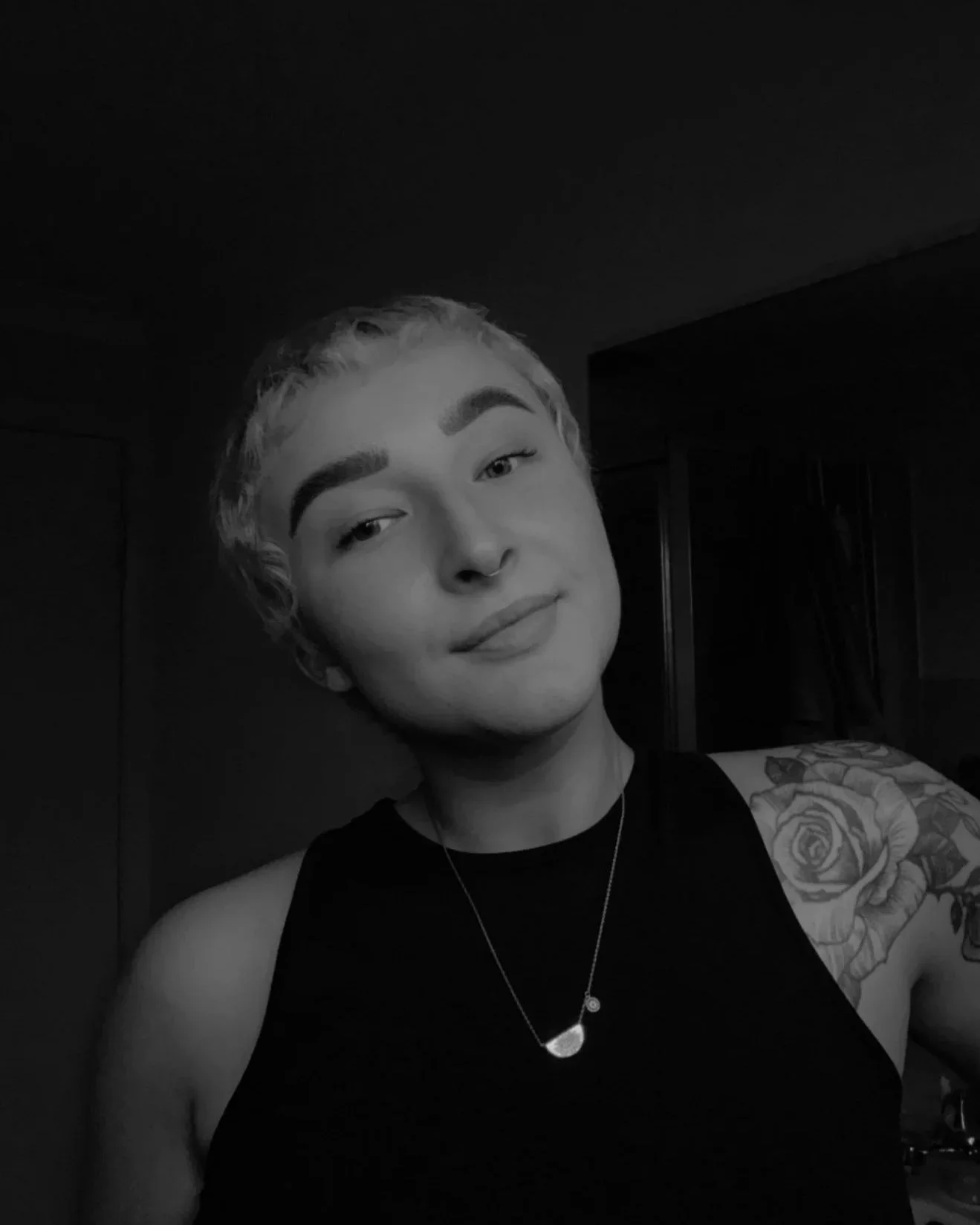 Black and white portrait of a young person with short, curly hair, wearing a sleeveless top, a necklace, and having a tattoo of roses on their right arm.
