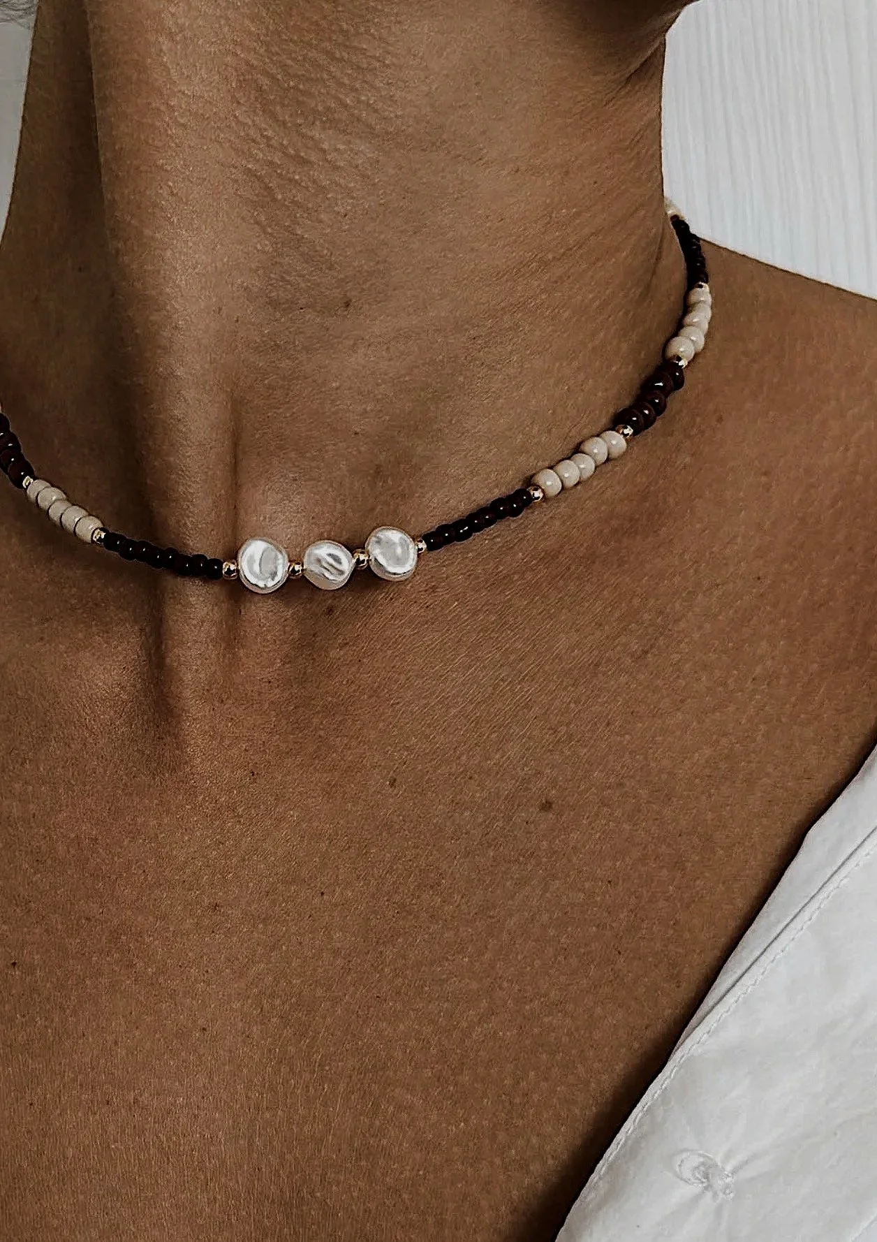 Close-up of a person's neck and shoulder wearing a beaded necklace with white pearl-like beads and small dark beads, and a white shirt.