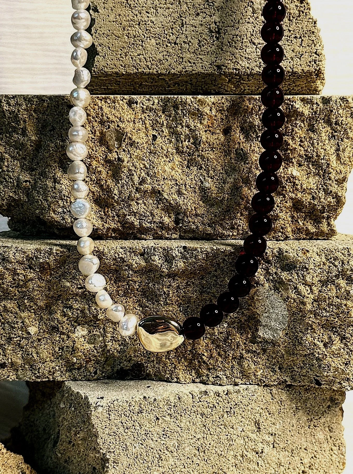 A necklace with white pearls on the left and dark red beads on the right, connected by a gold clasp, resting on stacked rough-textured stone blocks.