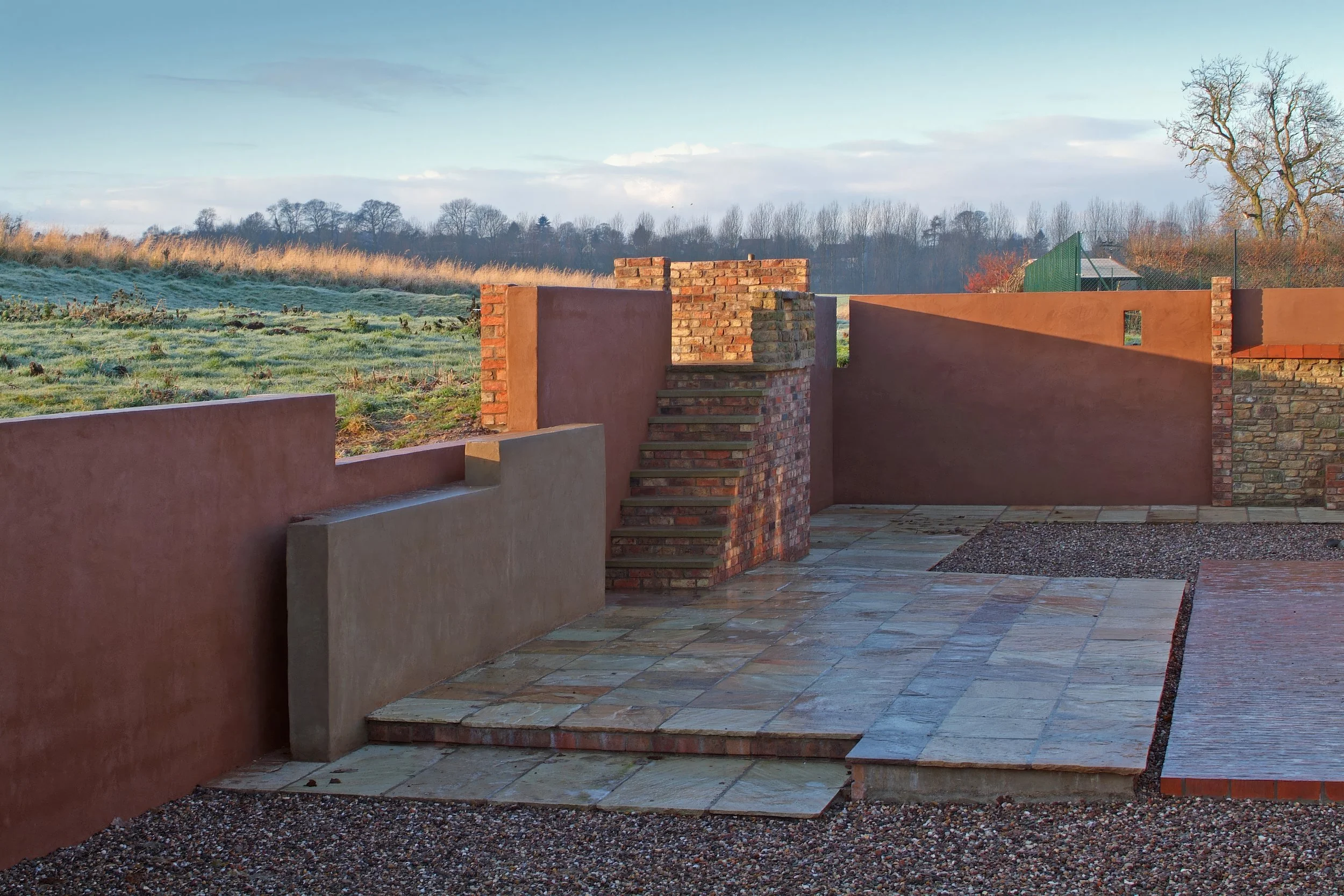 A partially constructed outdoor patio area with brick stairs, pink and brick walls, and a tiled ground with a scenic background of grassy fields, trees, and a partly cloudy sky.